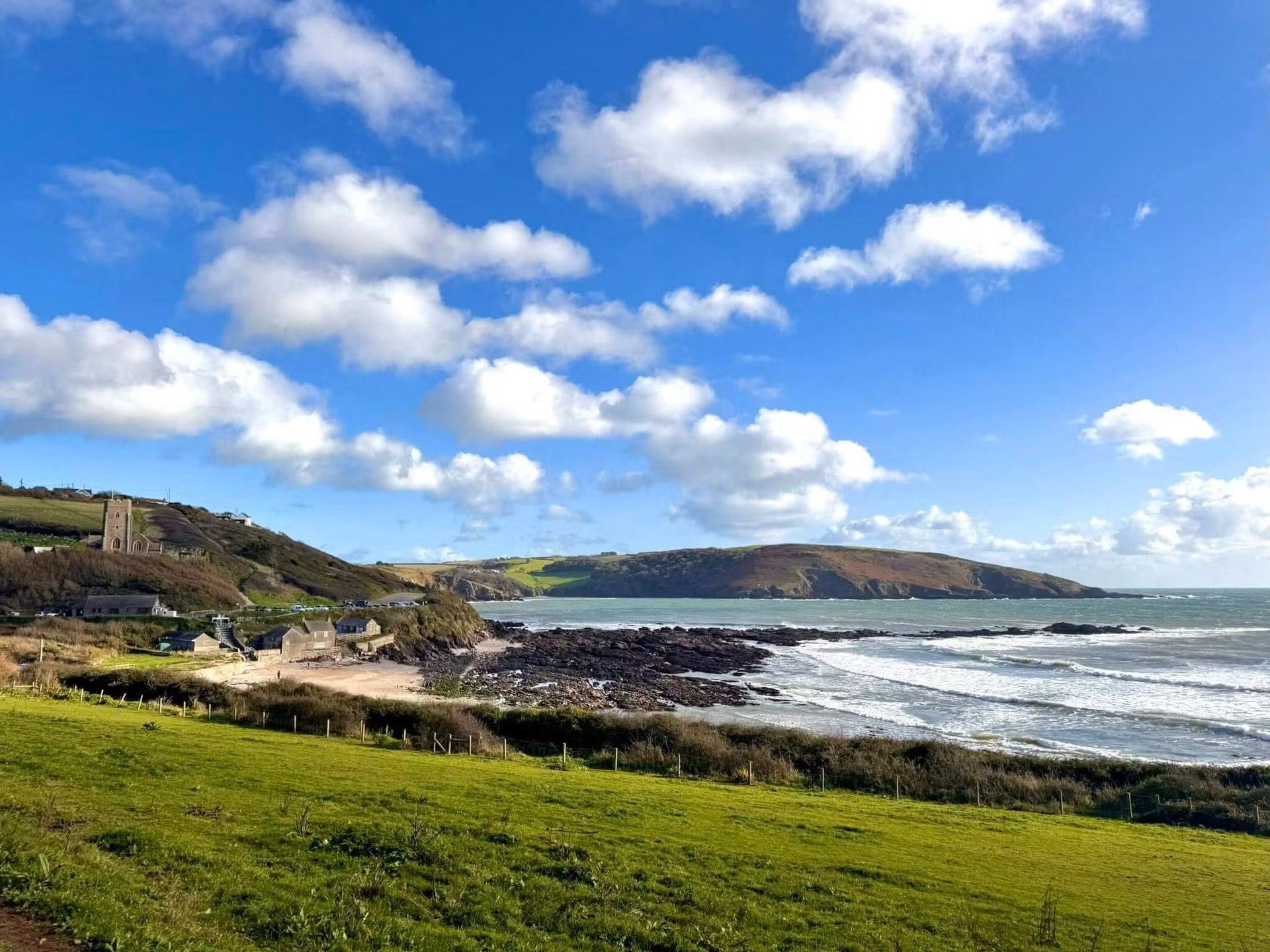 Coastal bay with rocky shoreline, green fields, and a small village under a blue sky with scattered clouds