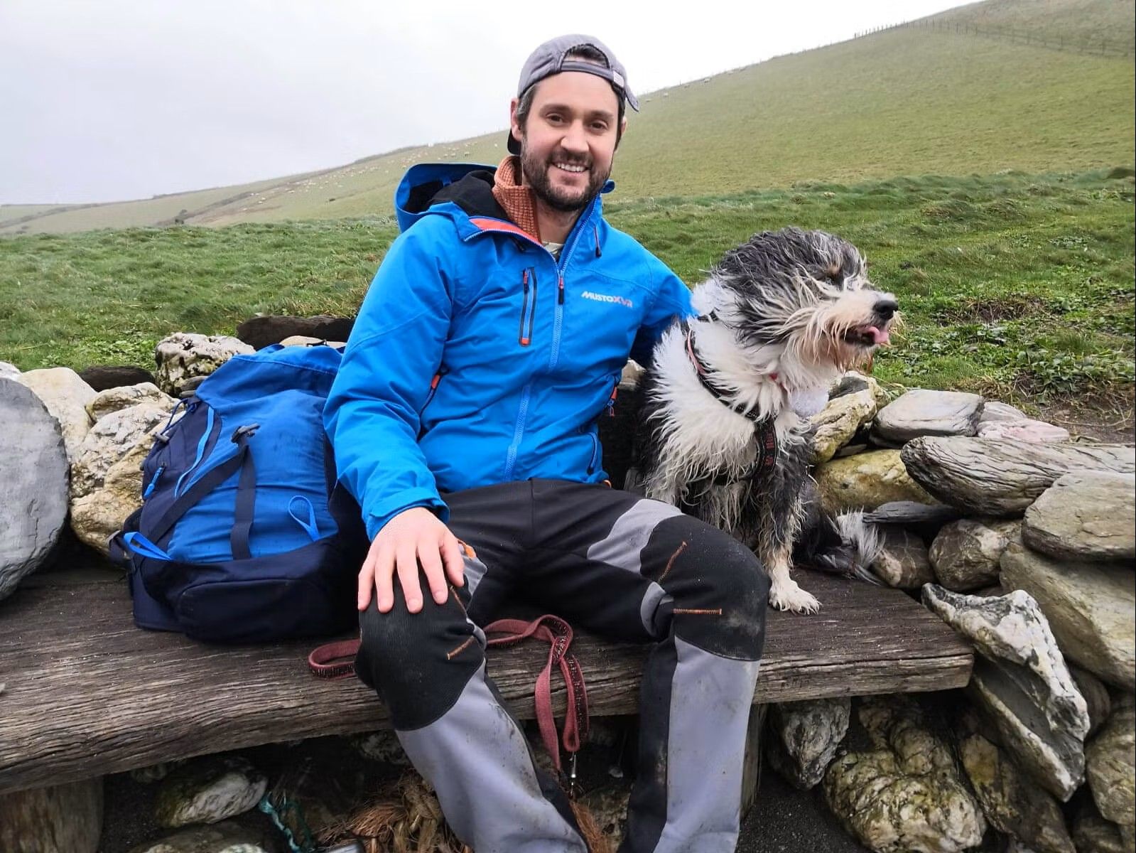 Hiker in a blue jacket sitting on a wooden bench with a shaggy black-and-white dog in a countryside setting