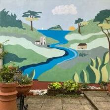 Painted mural of a winding blue river through green hills with a sunrise, trees and small buildings, viewed from a patio with plant pots.