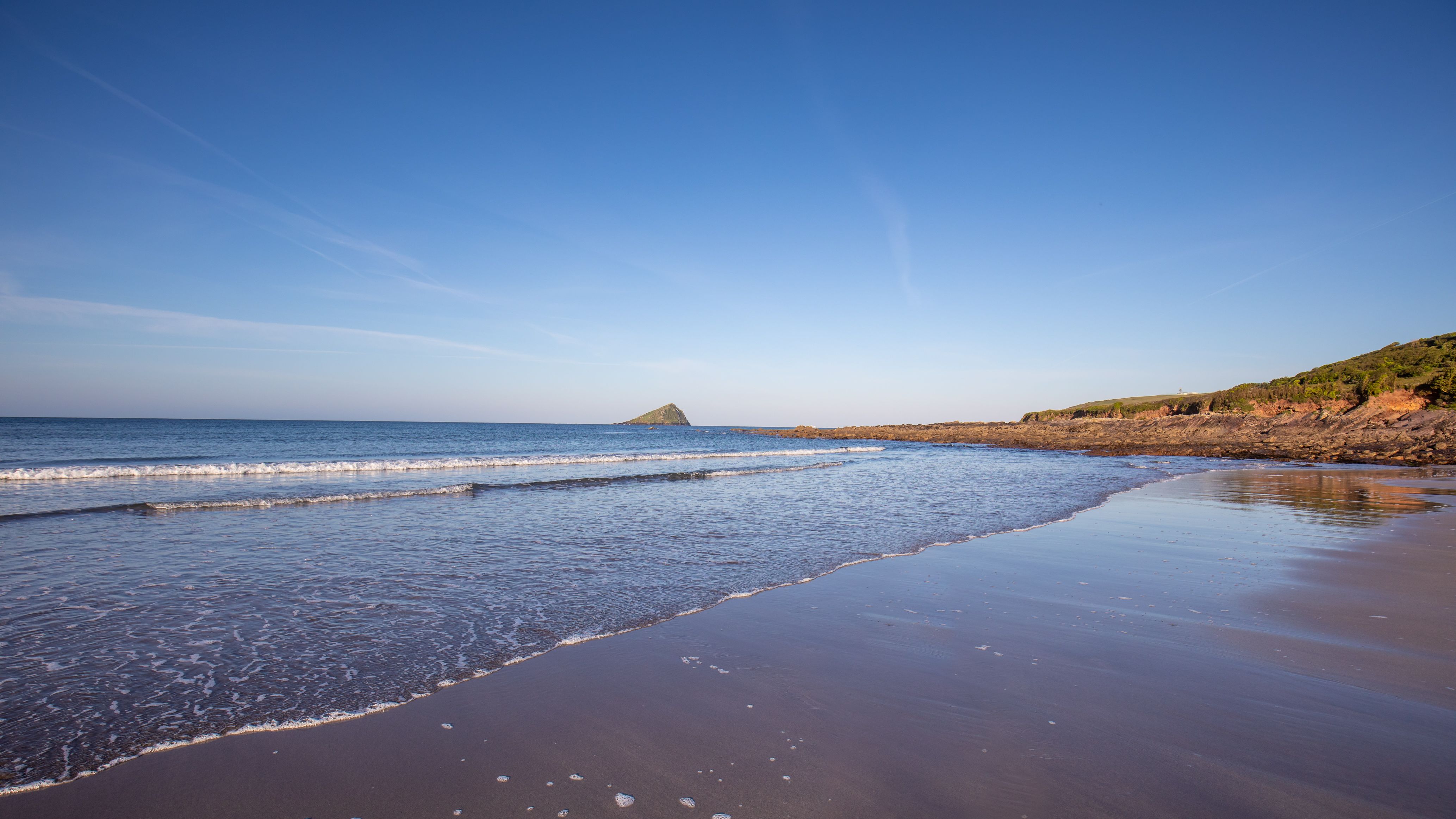 sandy beach with gentle waves, rocky shoreline, and a small island in the distance under a clear blue sky