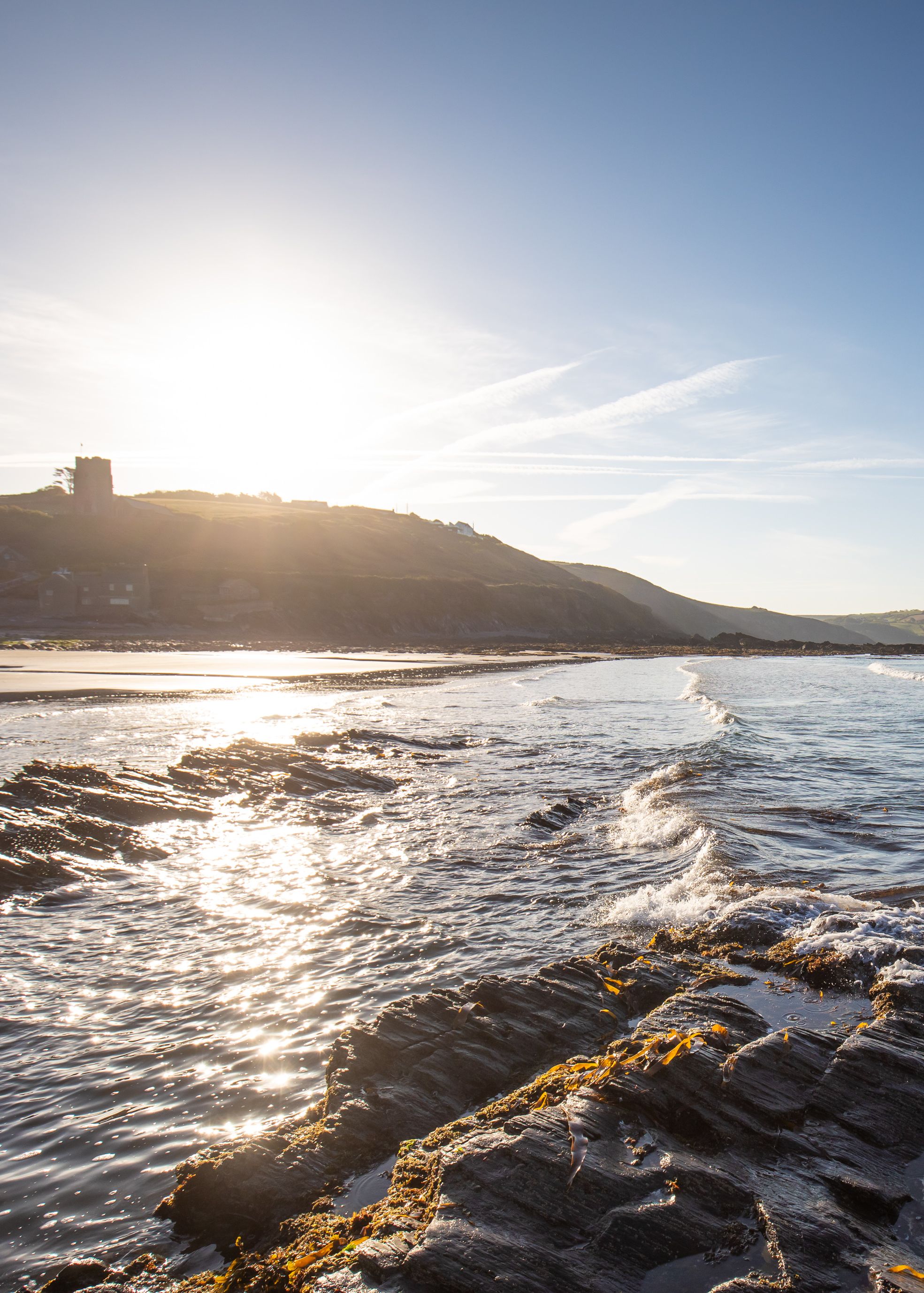 Rocky beach shoreline with gentle waves and hills in the background during sunset.