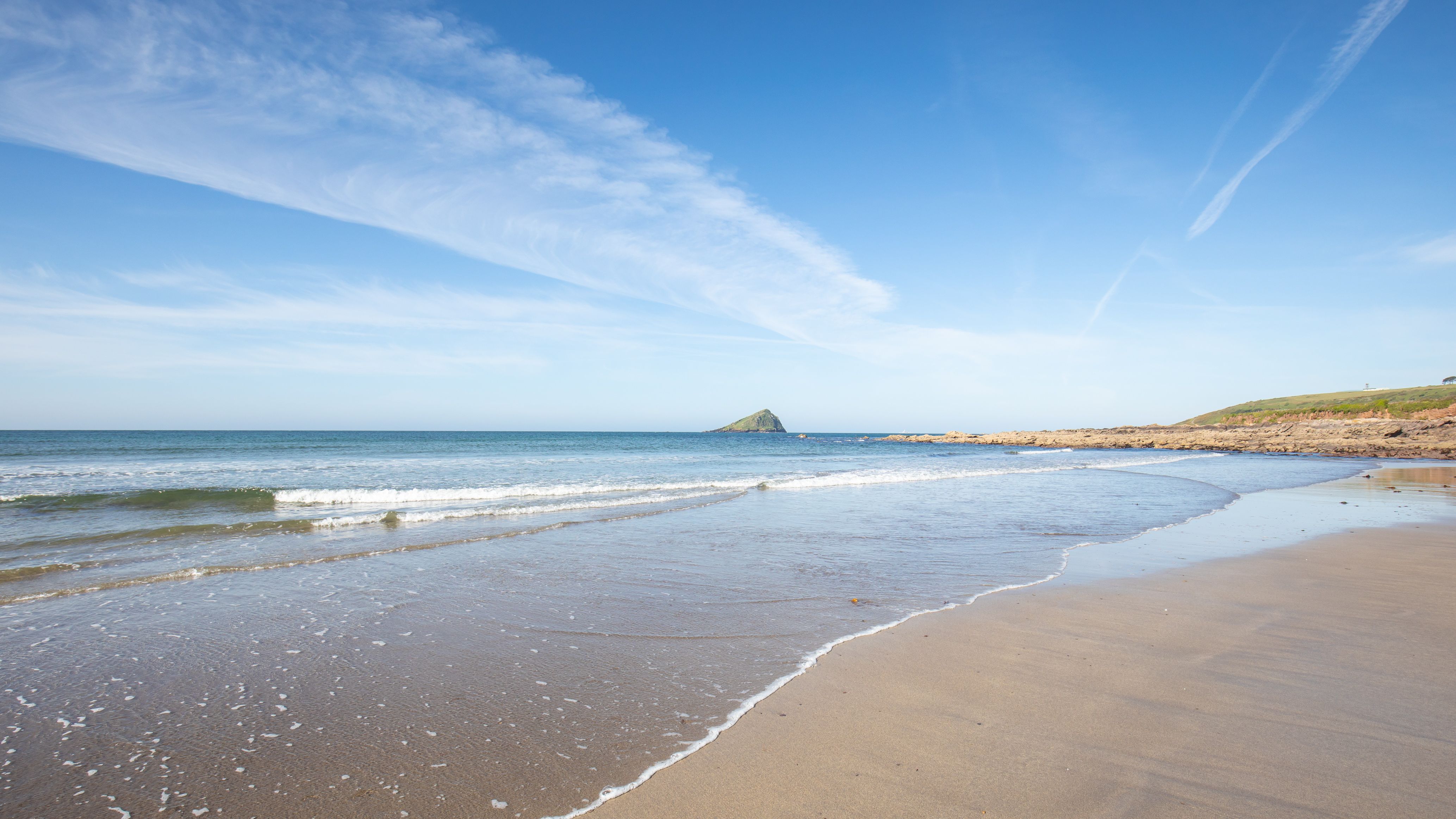 Wide sandy beach with gentle waves under a clear blue sky, an island visible on the horizon.