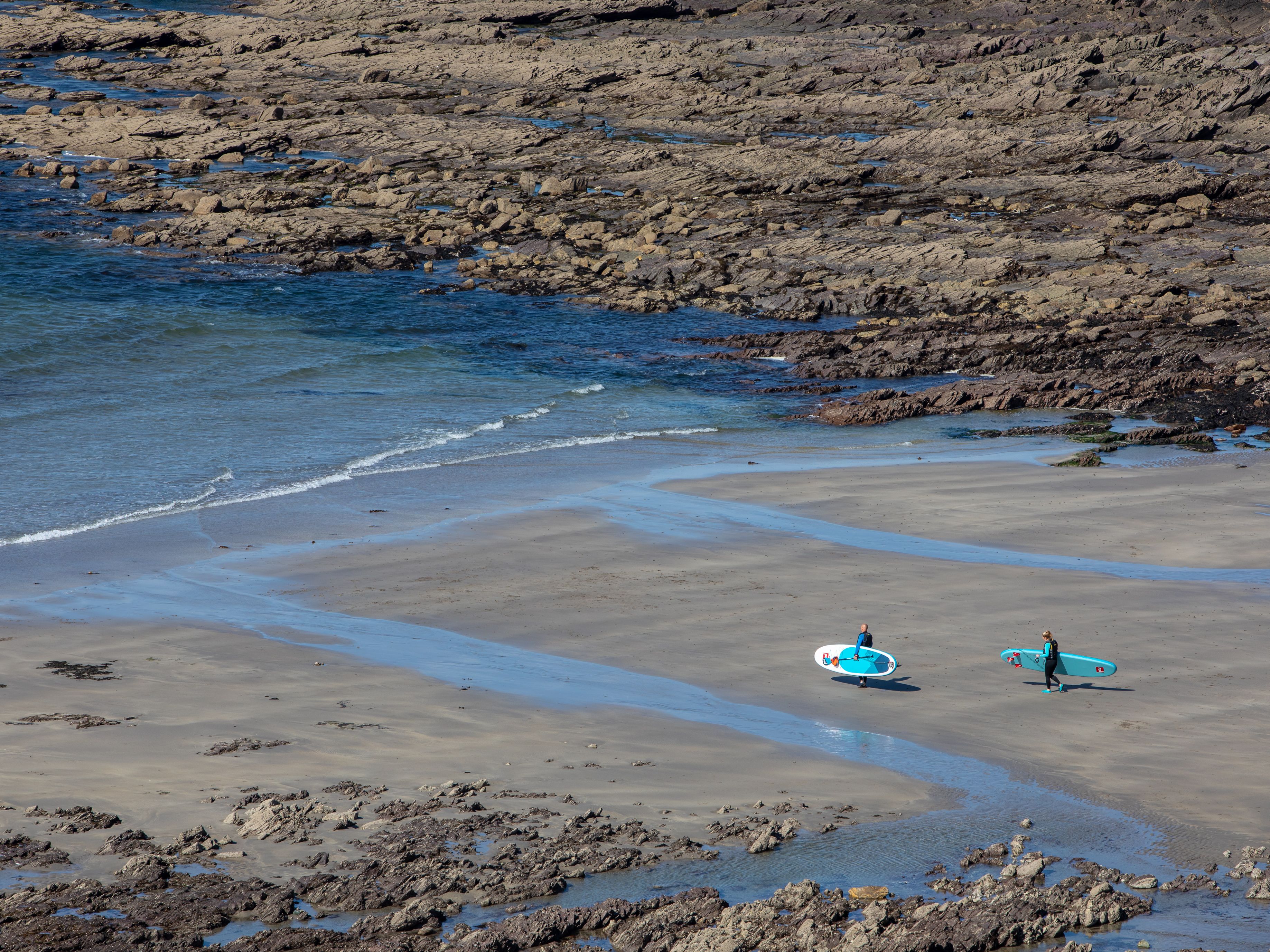 Two people carrying surfboards on a rocky beach with waves approaching the shore