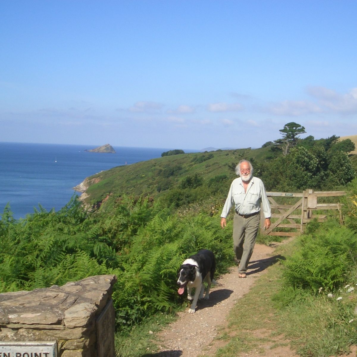 Man walking with a dog along a coastal path, with the sea and greenery in the background.