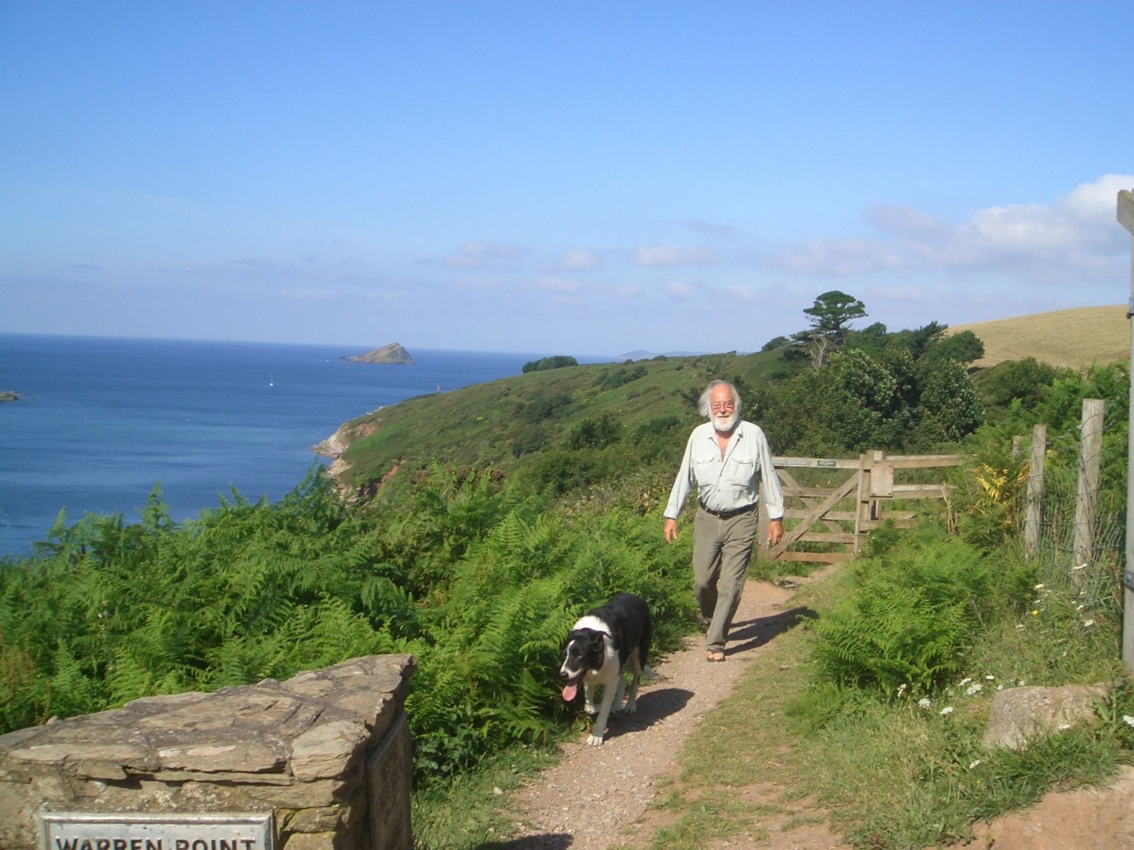 Man walking with a dog along a coastal path, with the sea and greenery in the background.