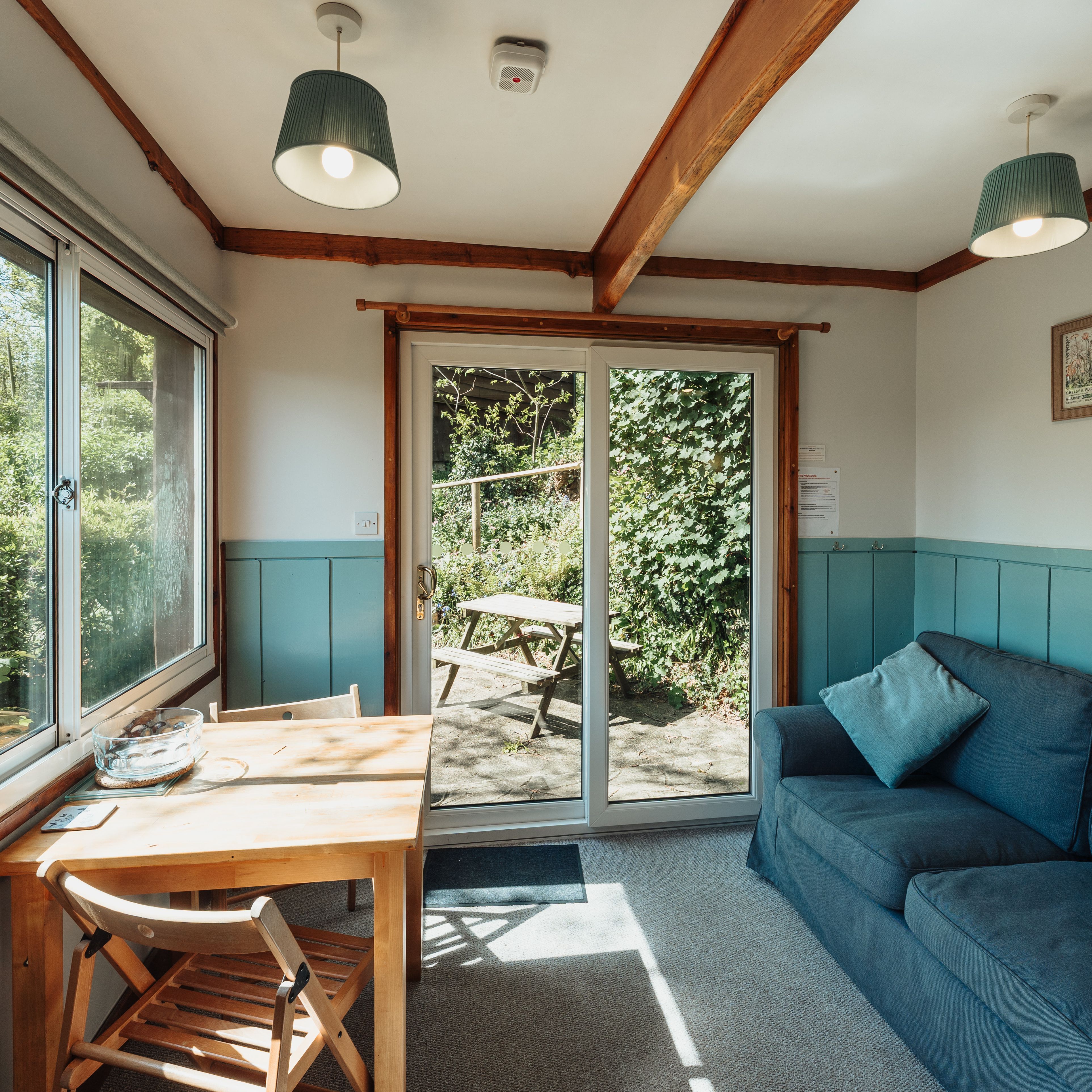 Cosy sitting room with blue sofa, wooden table and large windows overlooking woodland, with French doors to a patio picnic bench.