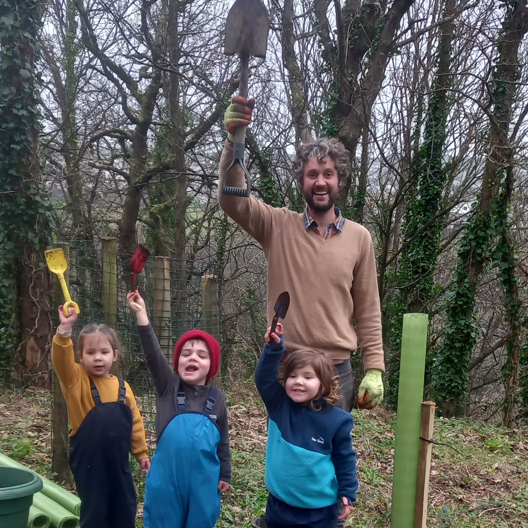 Adult and three children holding up garden spades in a woodland planting area beside a pink tub of compost