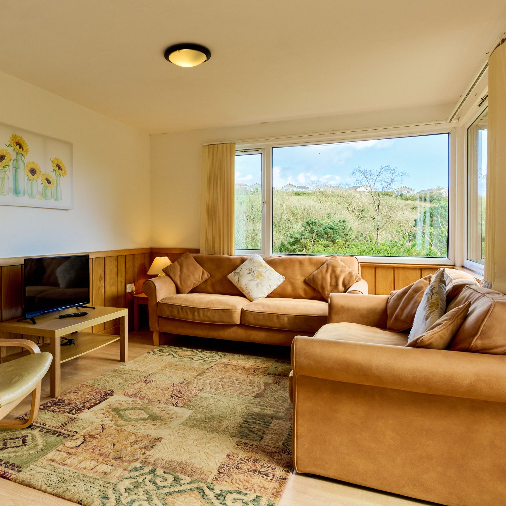 Cozy living room with two tan sofas, an armchair, a patterned rug, TV, and large windows with countryside views.
