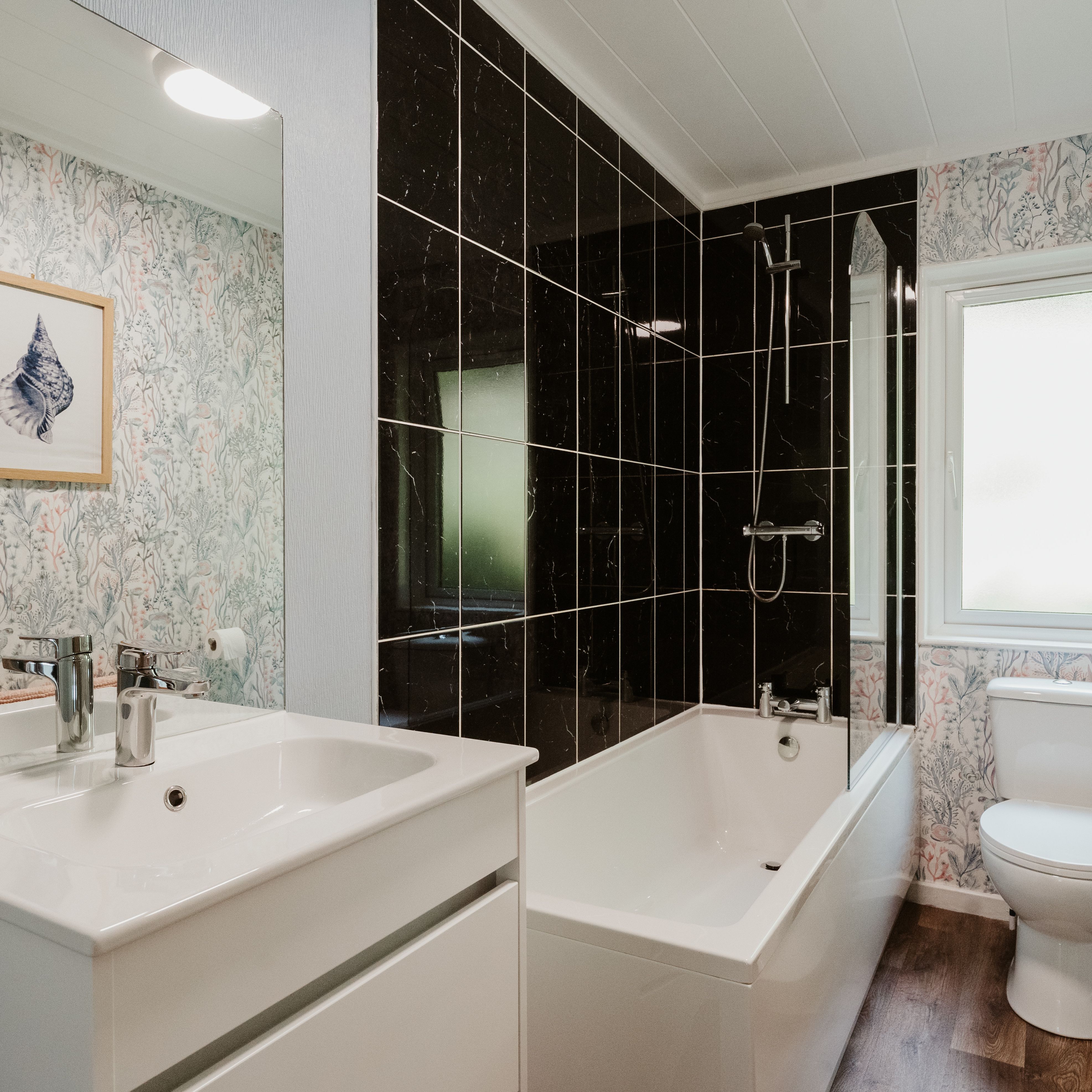 Bright bathroom with white vanity, bath and shower, black wall tiles and floral wallpaper