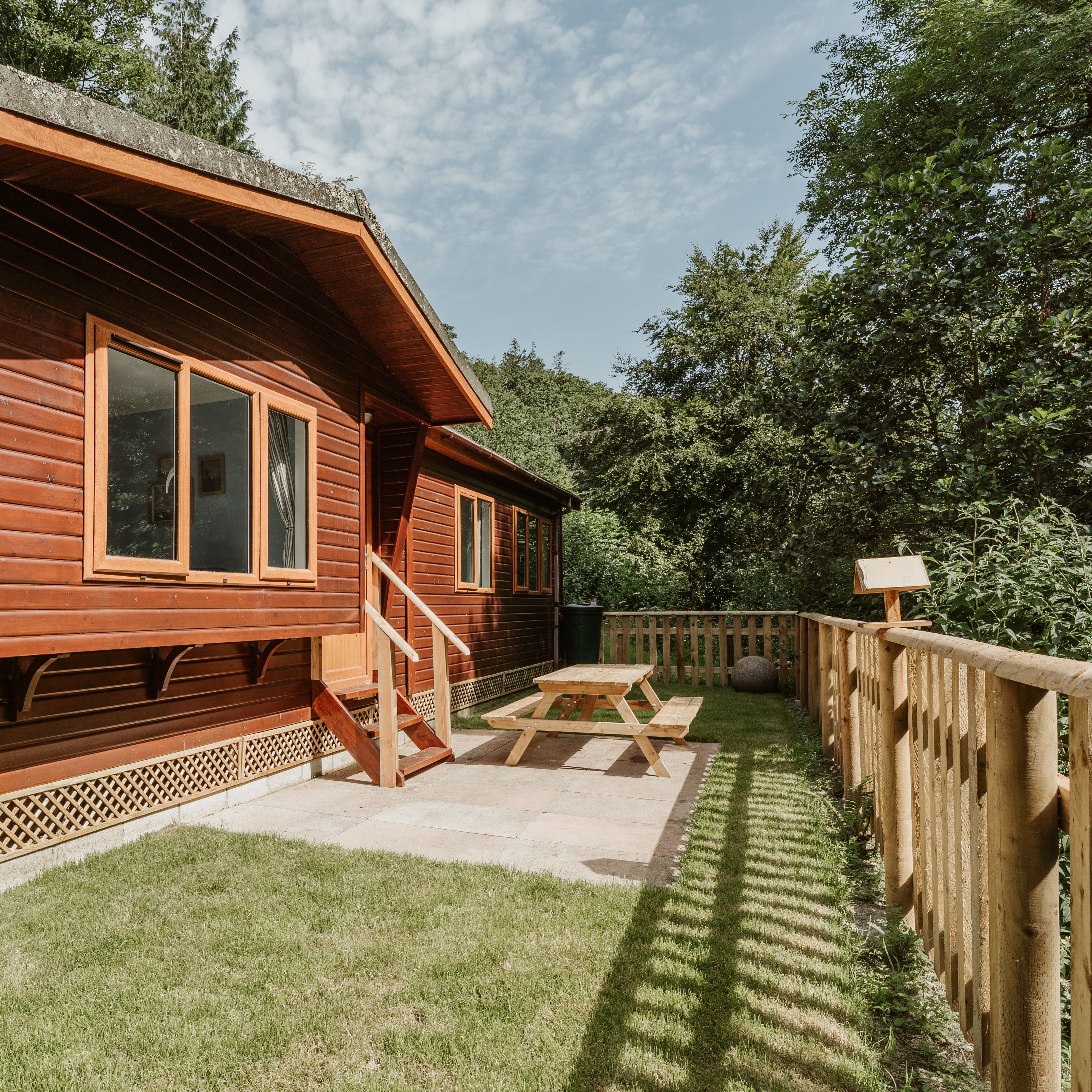 Wooden lodge exterior with fenced patio and picnic table in a leafy setting