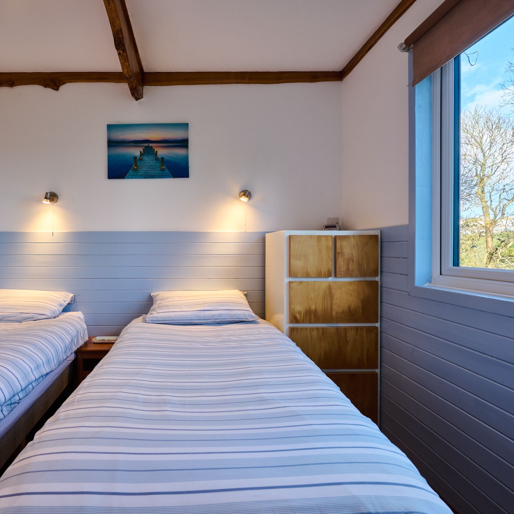 Twin bedroom with two single beds, striped bedding, a window, and a painting of a dock over water on the wall.