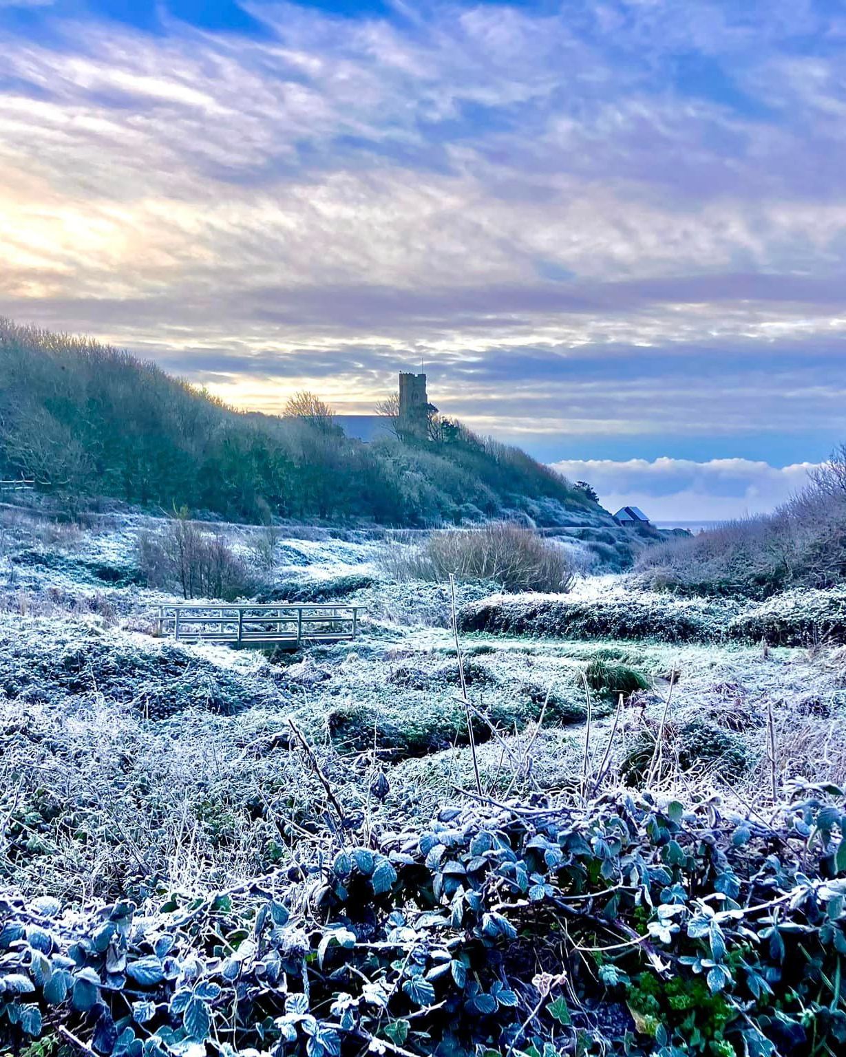 Frost-covered coastal valley at sunrise with a small footbridge and a distant stone tower