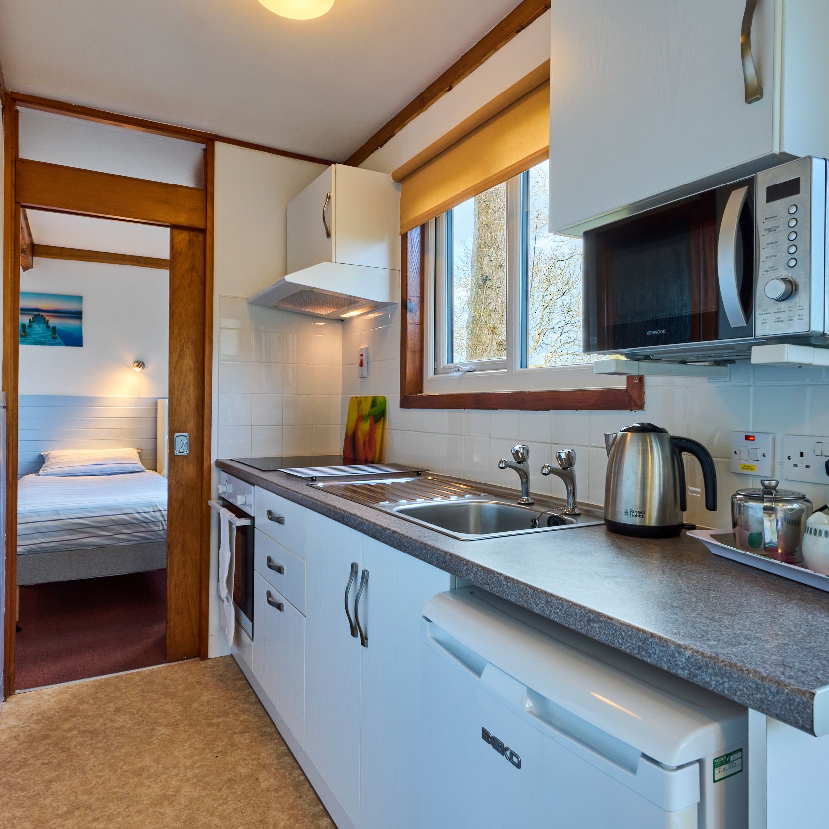 Small kitchen with white cabinets, microwave, kettle and view of a bedroom through open doorway