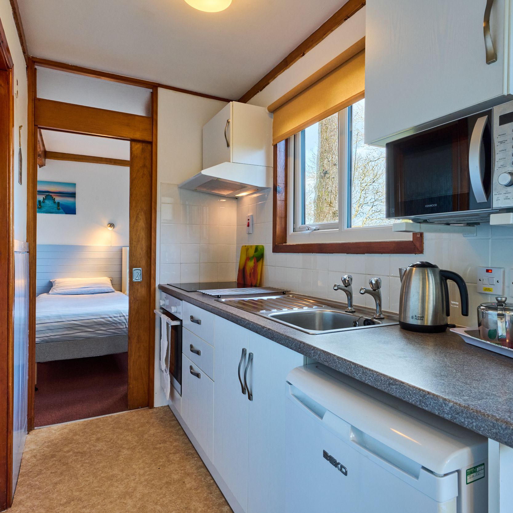 Small kitchen with white cabinets, microwave, kettle and view of a bedroom through open doorway