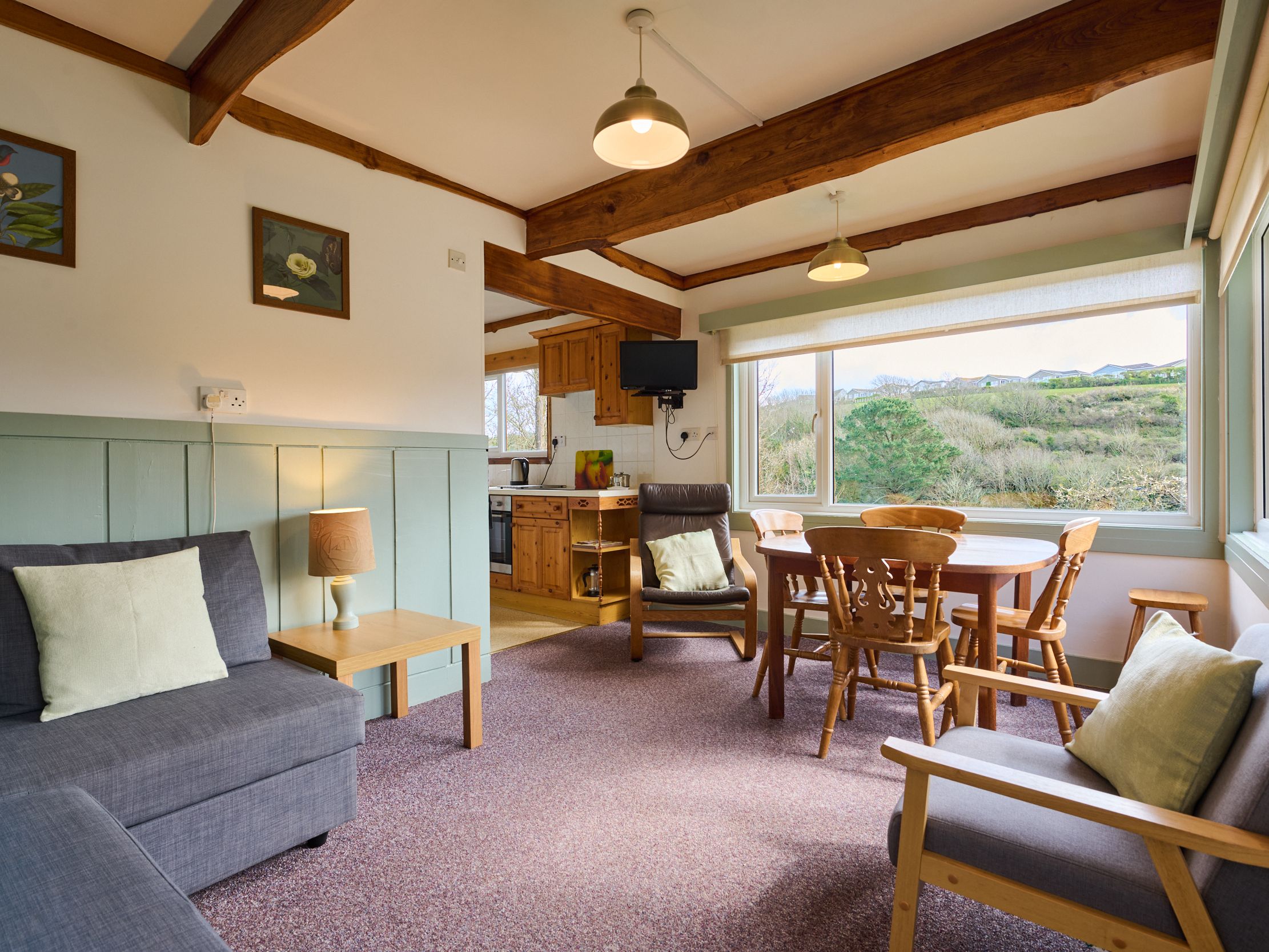 Bright living and dining area with large window overseeing green hills, featuring wooden beams, a round dining table, armchairs, and a modern sofa.
