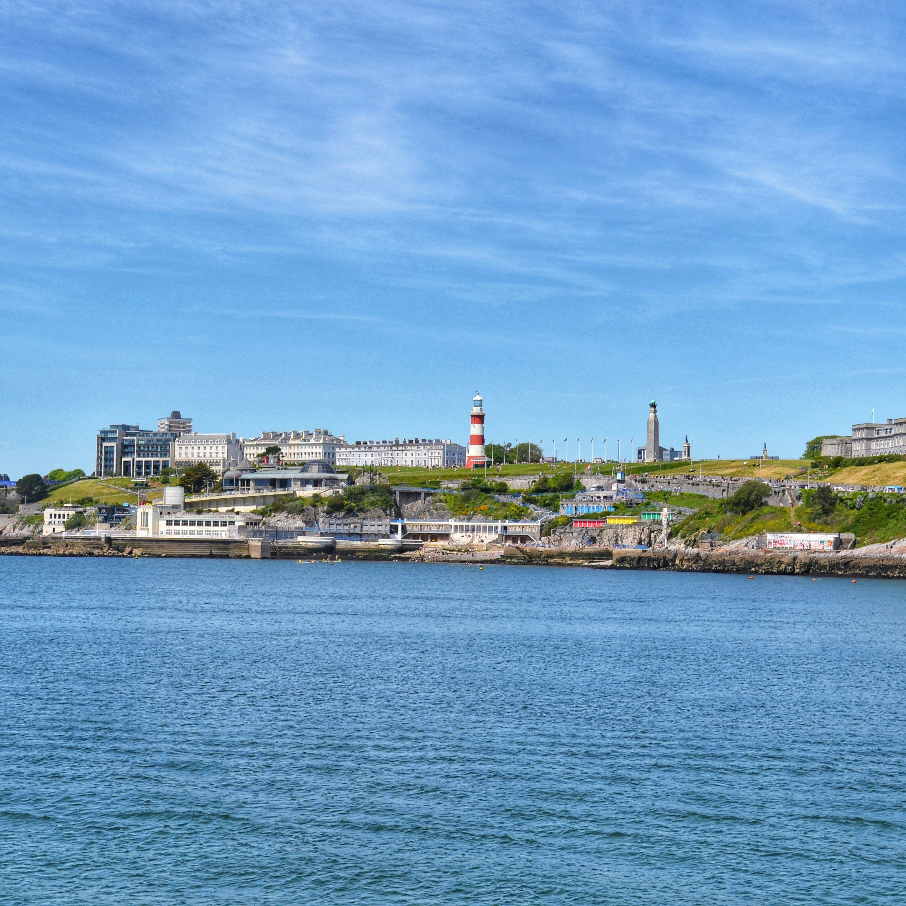 View of a coastal city with a lighthouse, historic buildings, and calm blue water under a clear sky.