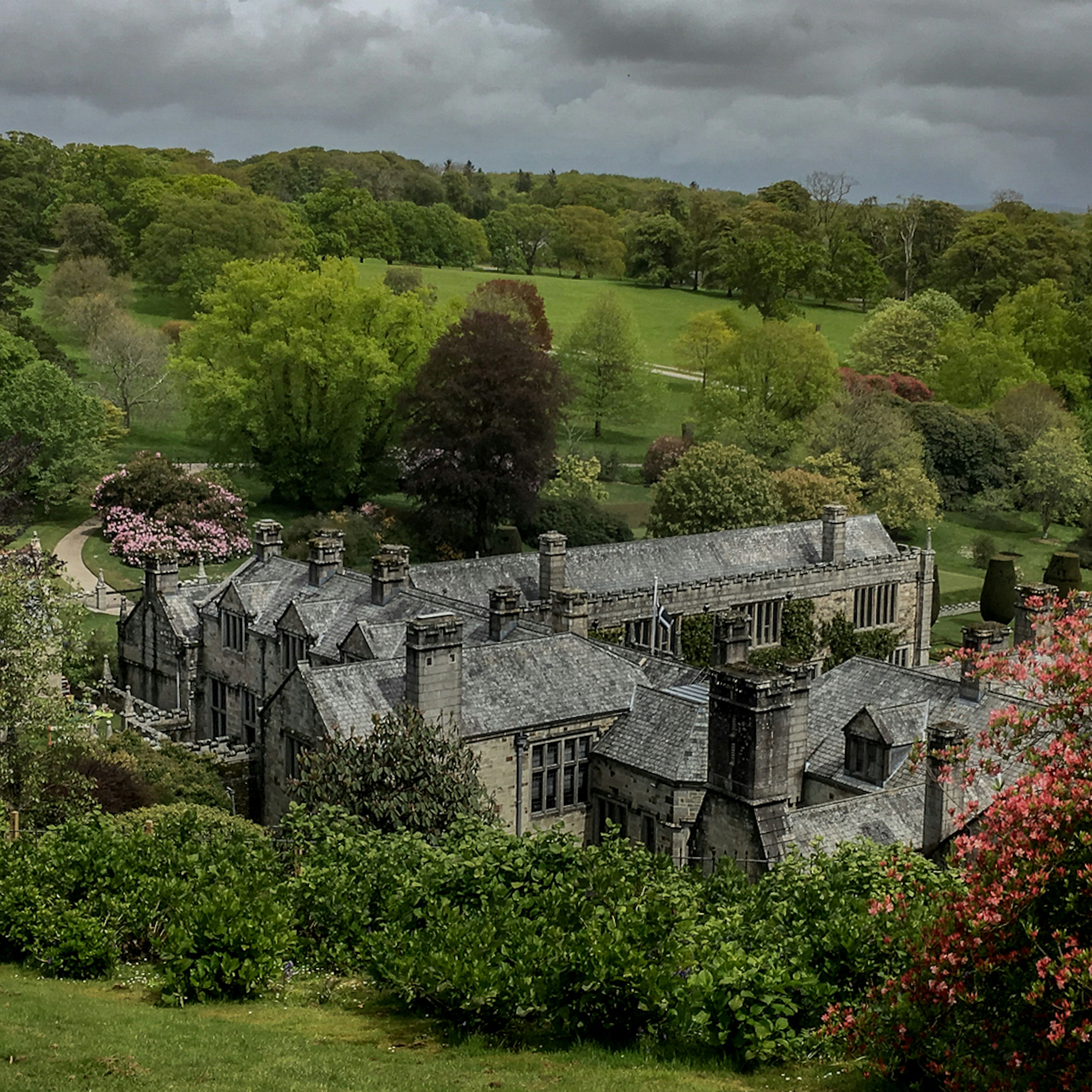 Historic stone manor house surrounded by lush greenery and flowering bushes under a cloudy sky.