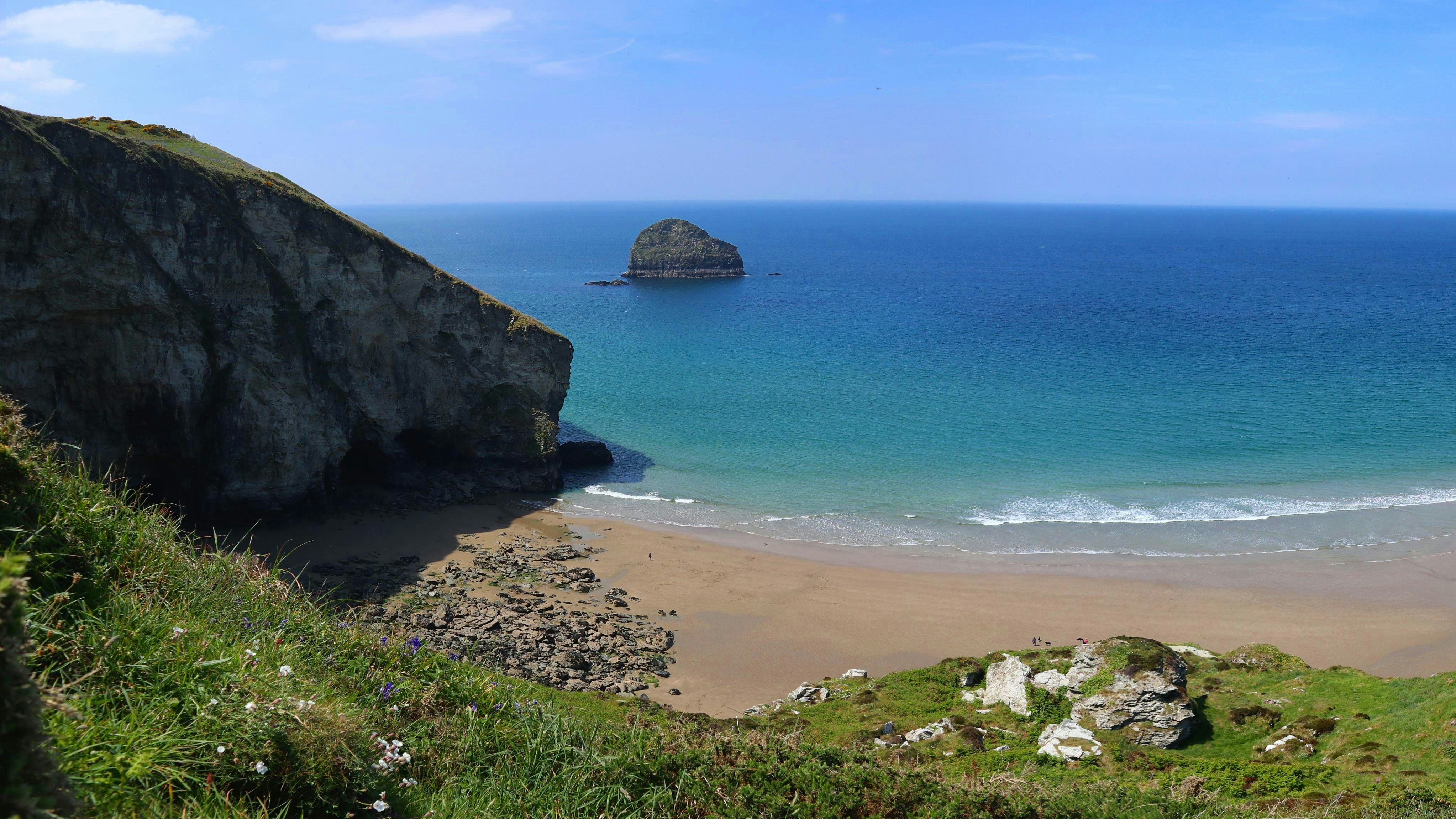 A panoramic view of a sandy beach with clear blue waters, surrounded by rocky cliffs and green hills.
