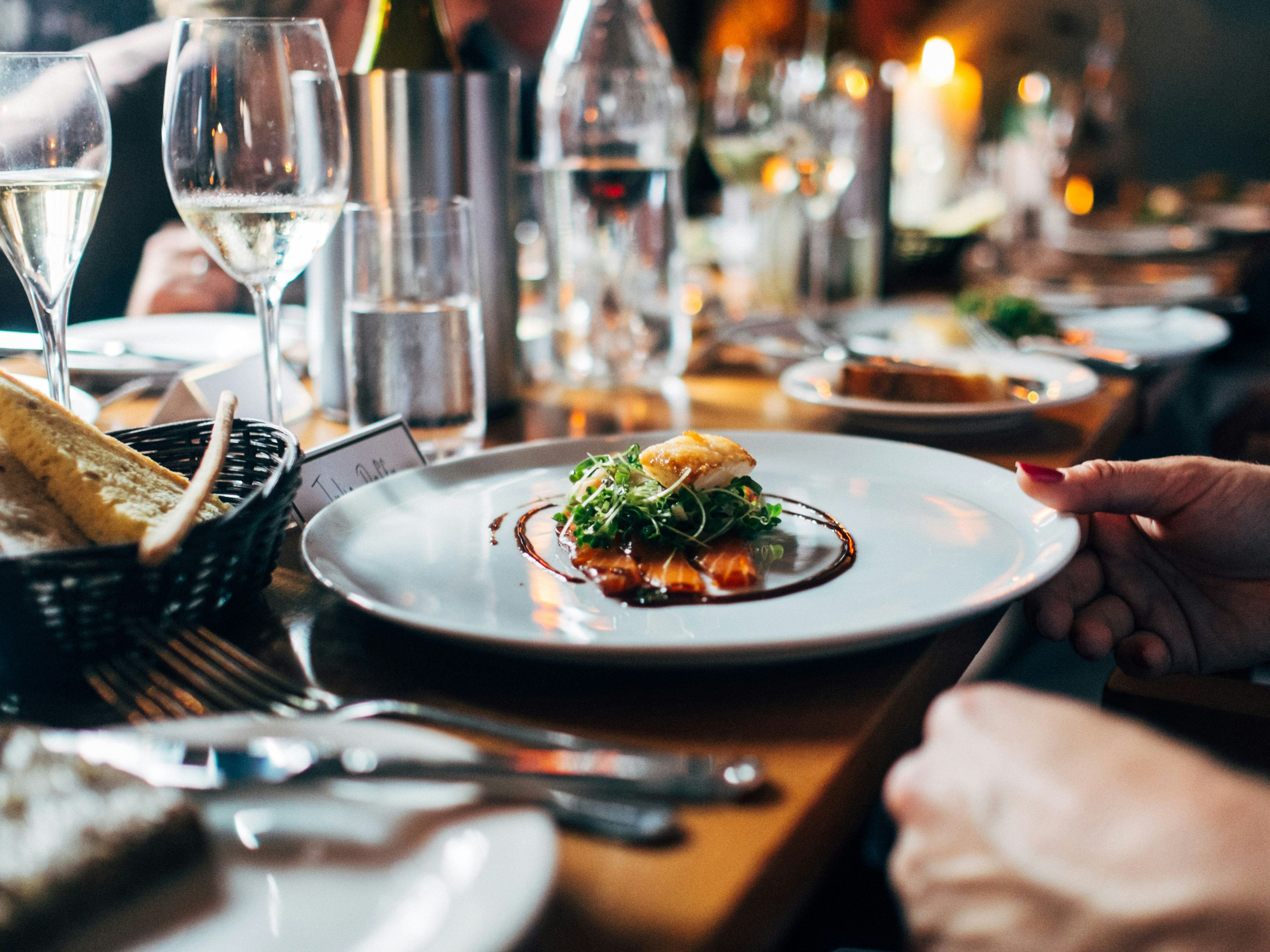 A plated gourmet dish with greens and sauce, set on a restaurant table with wine glasses and bread.