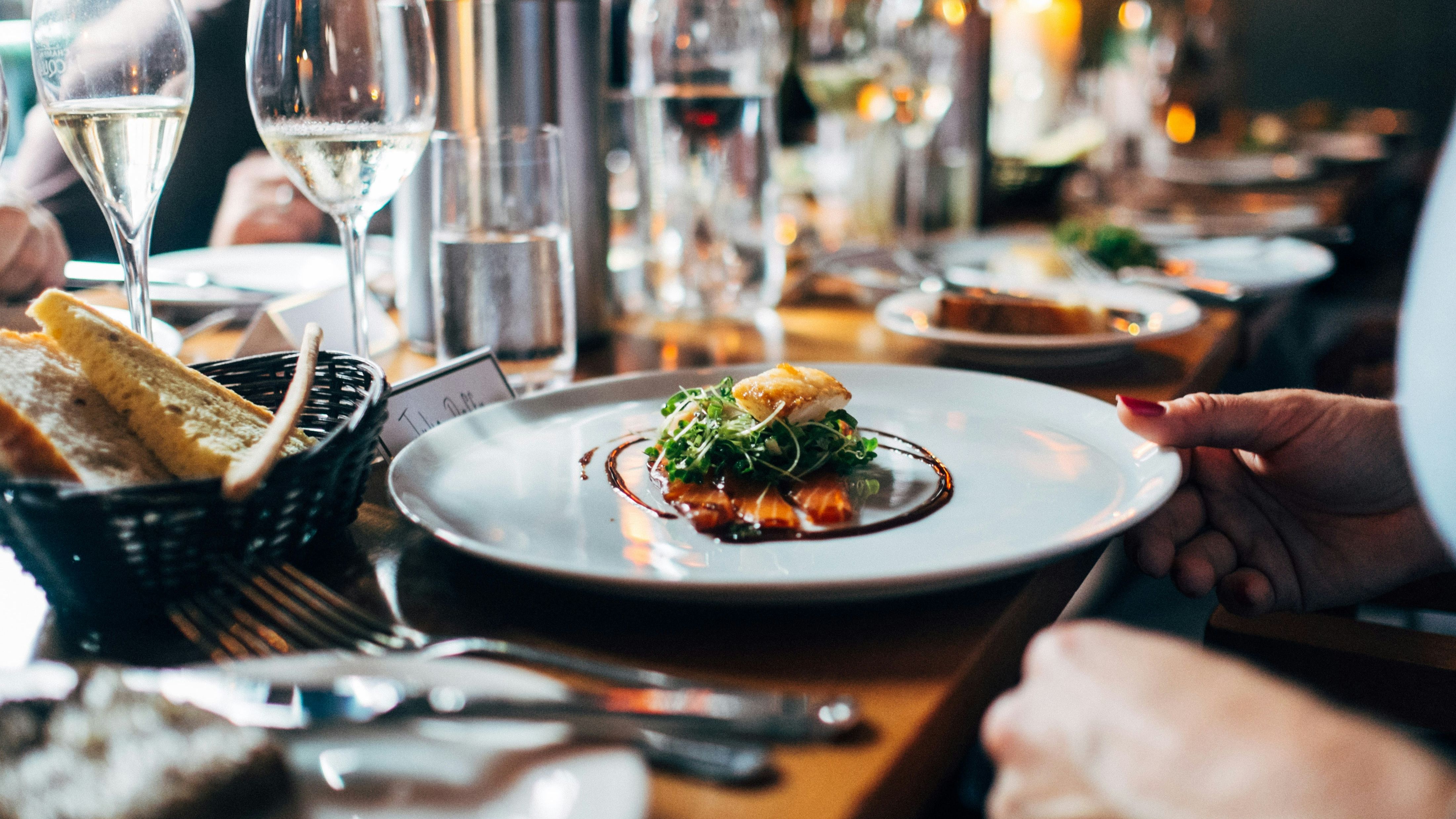 A plated gourmet dish with greens and sauce, set on a restaurant table with wine glasses and bread.