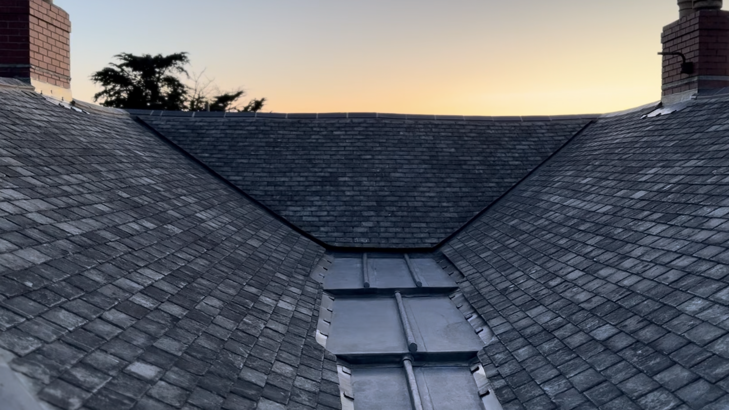 A view of a slate tiled roof at sunset with two brick chimneys on either side.