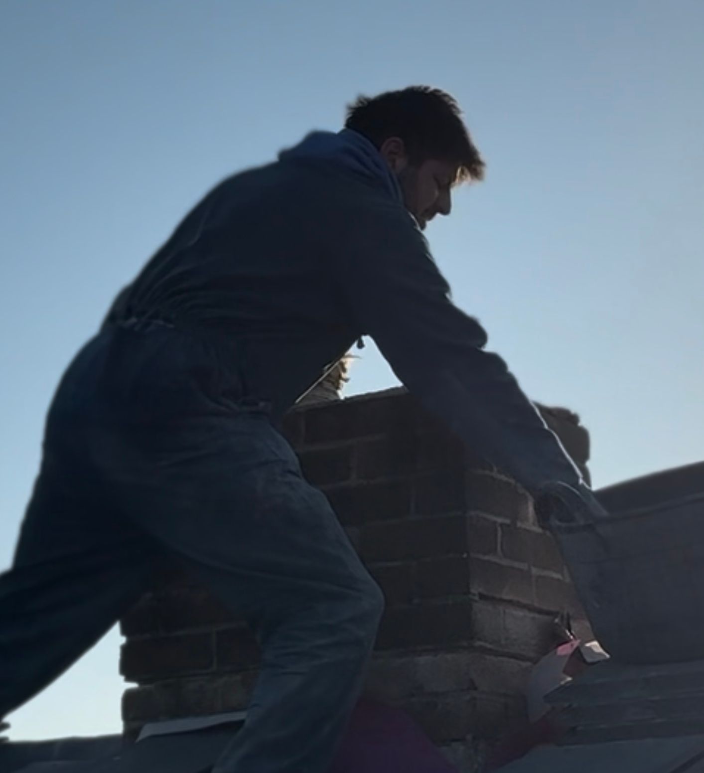 Person in work attire on a roof, placing a bucket near a brick chimney.