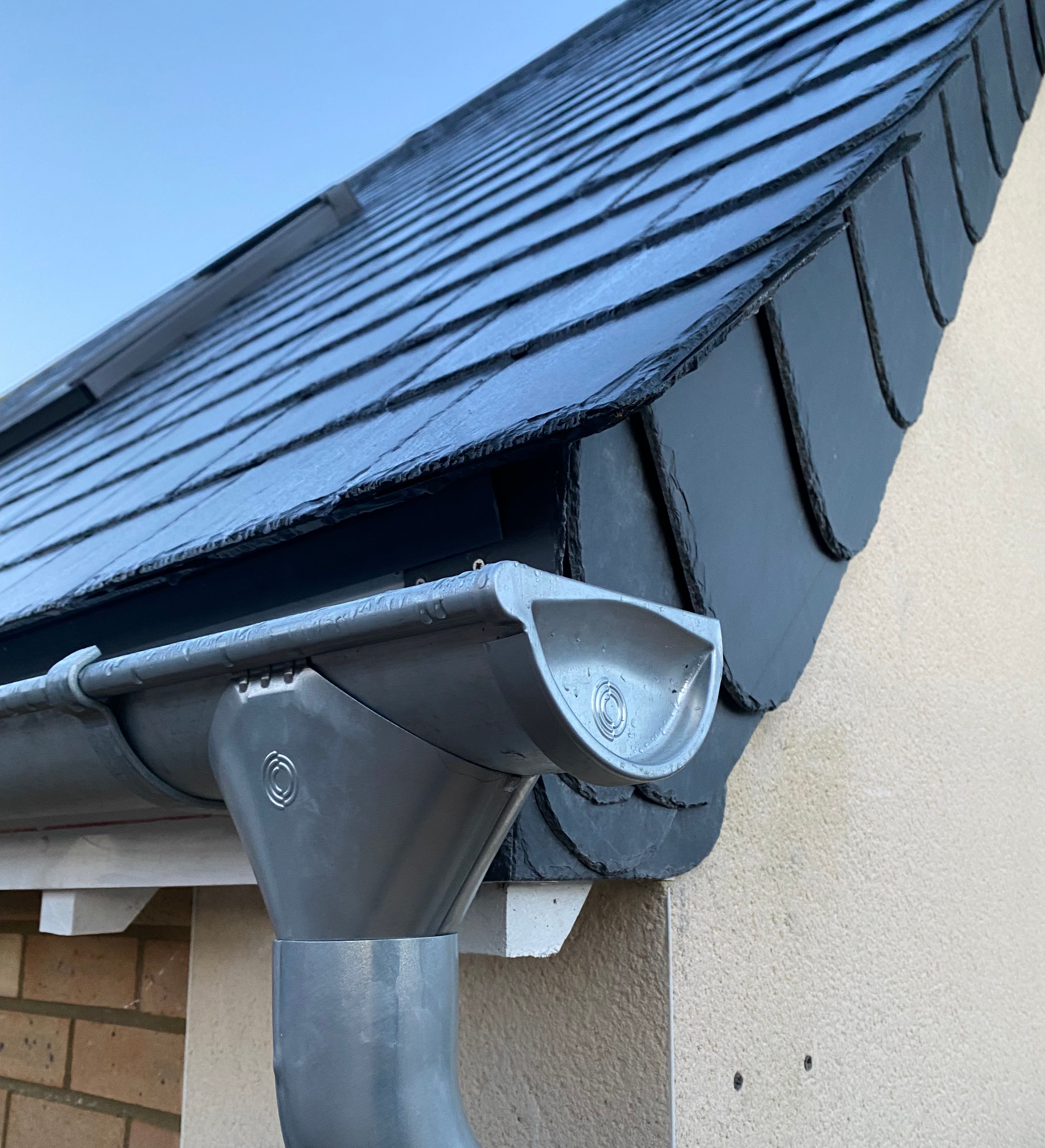 Close-up view of a grey plastic gutter and downpipe system attached to the corner of a house roof.