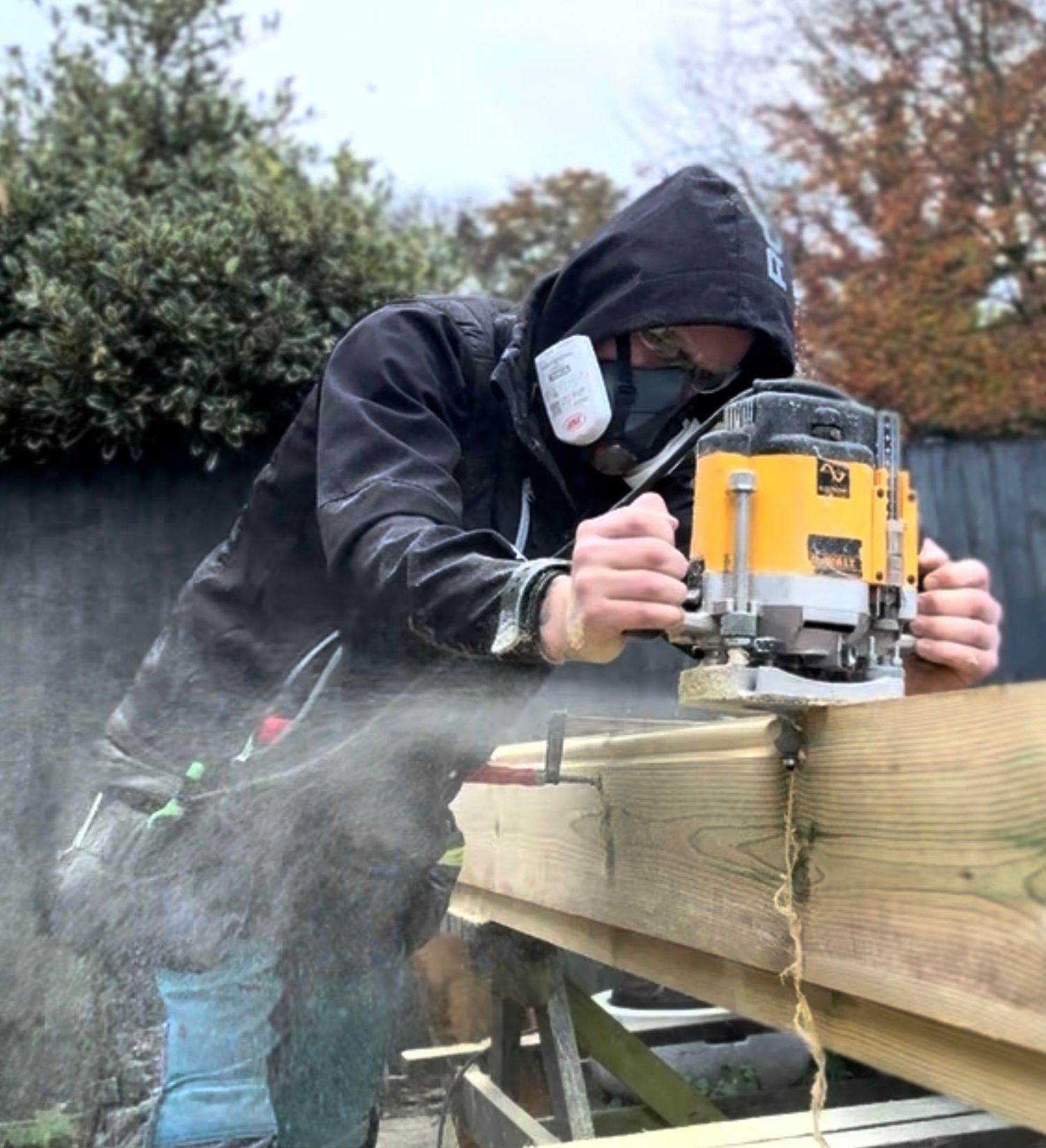 Person using a yellow router tool to shape a wooden beam outdoors, wearing safety goggles and a respirator.