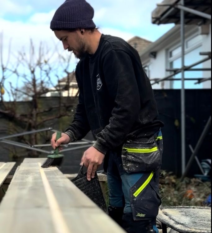 Person painting long wooden planks outdoors, wearing a beanie and work trousers.