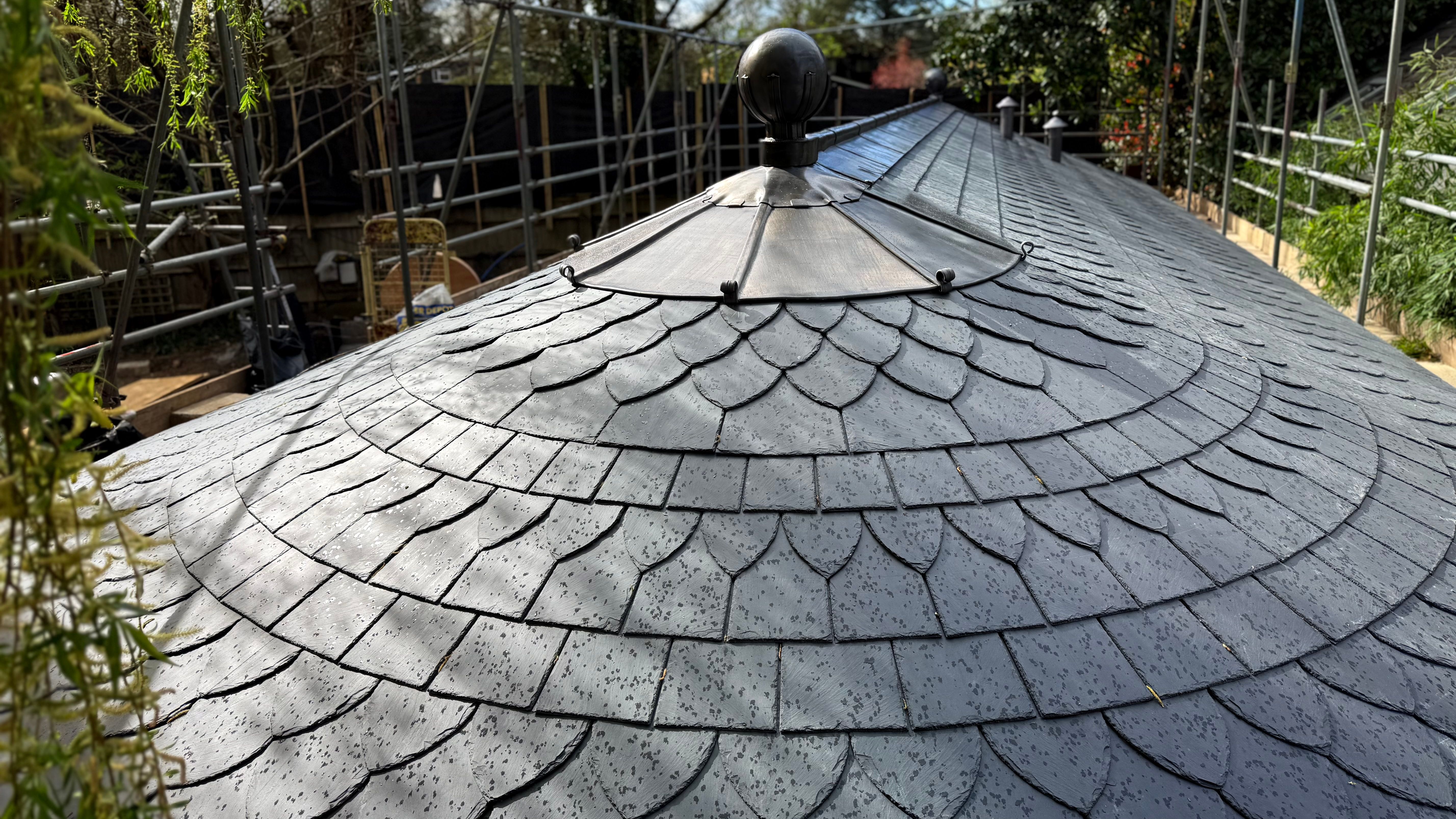 Curved slate roof with metal detailing and raindrops, surrounded by scaffolding and trees.
