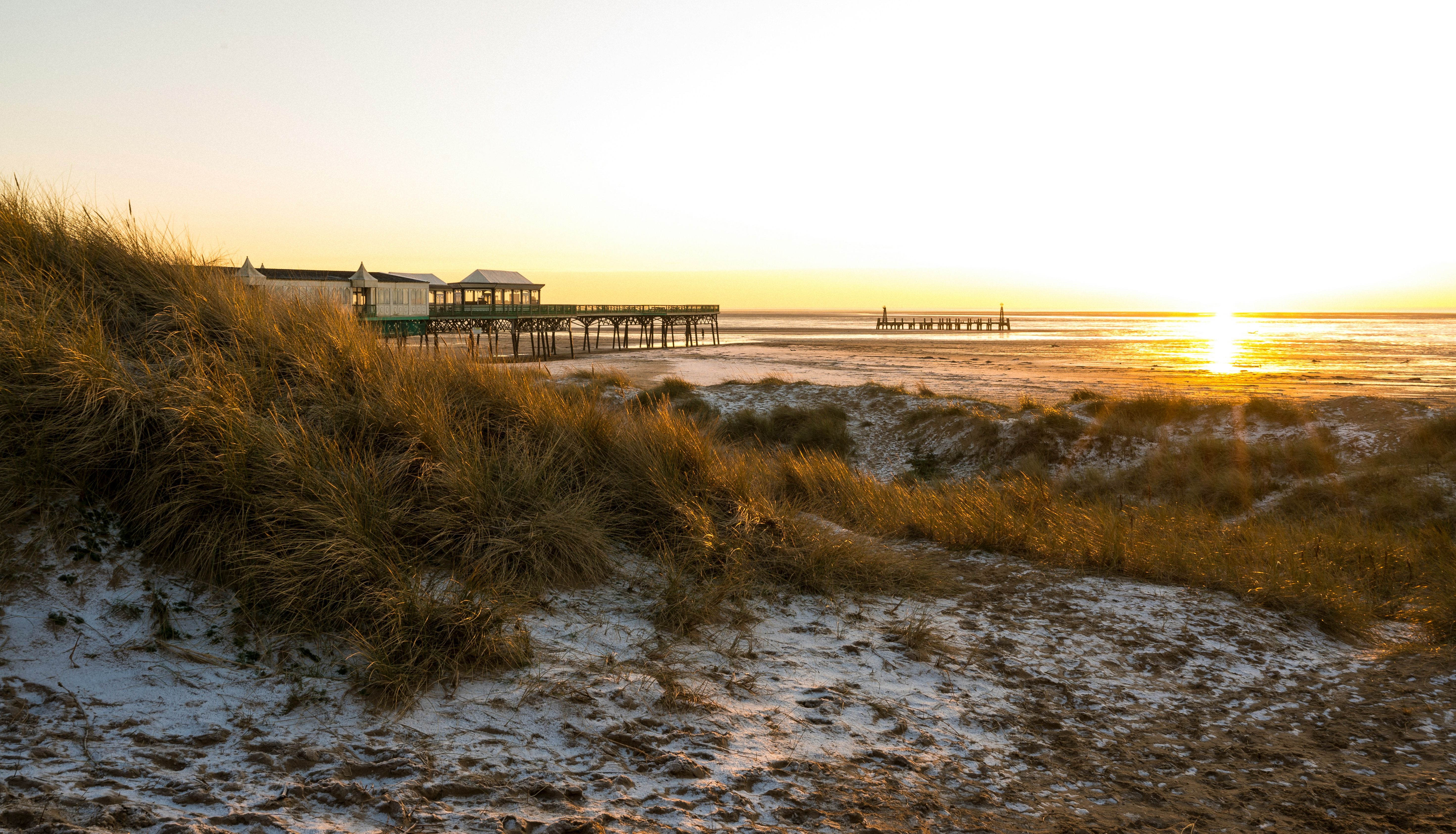 Sunset over a sandy beach with dune grass and a pier stretching into the sea