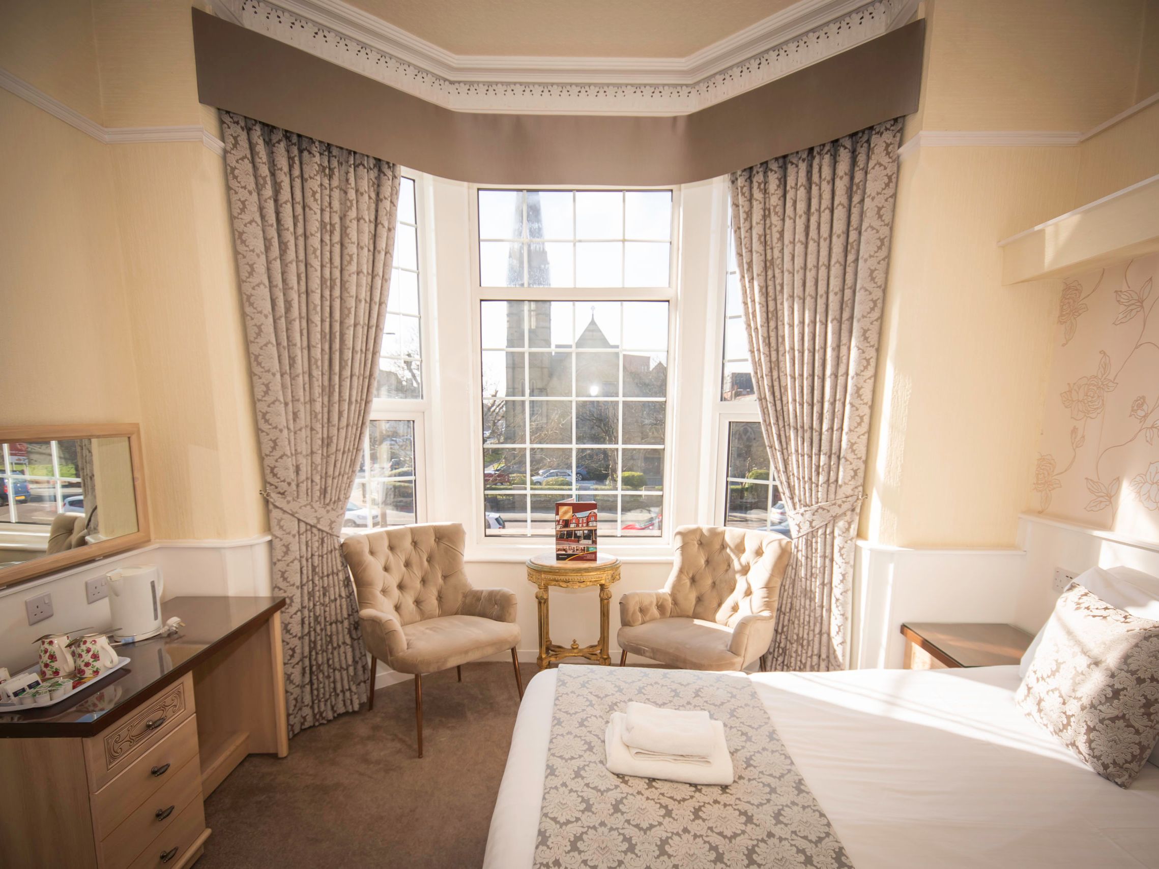 Sunlit hotel bedroom with large bay window, patterned curtains, two armchairs and neatly made bed