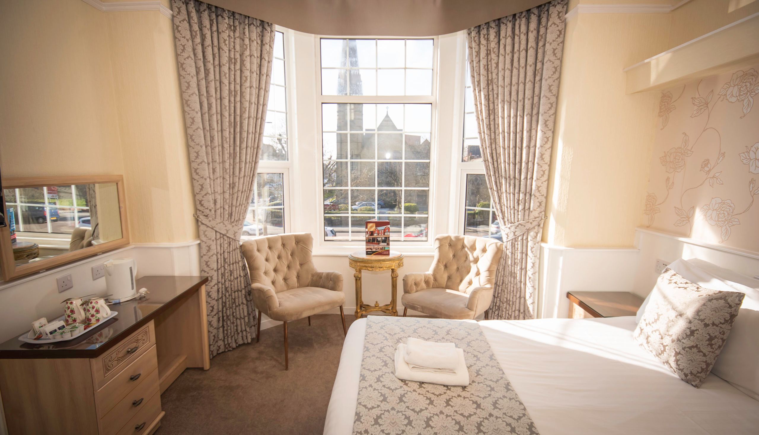 Sunlit hotel bedroom with large bay window, patterned curtains, two armchairs and neatly made bed