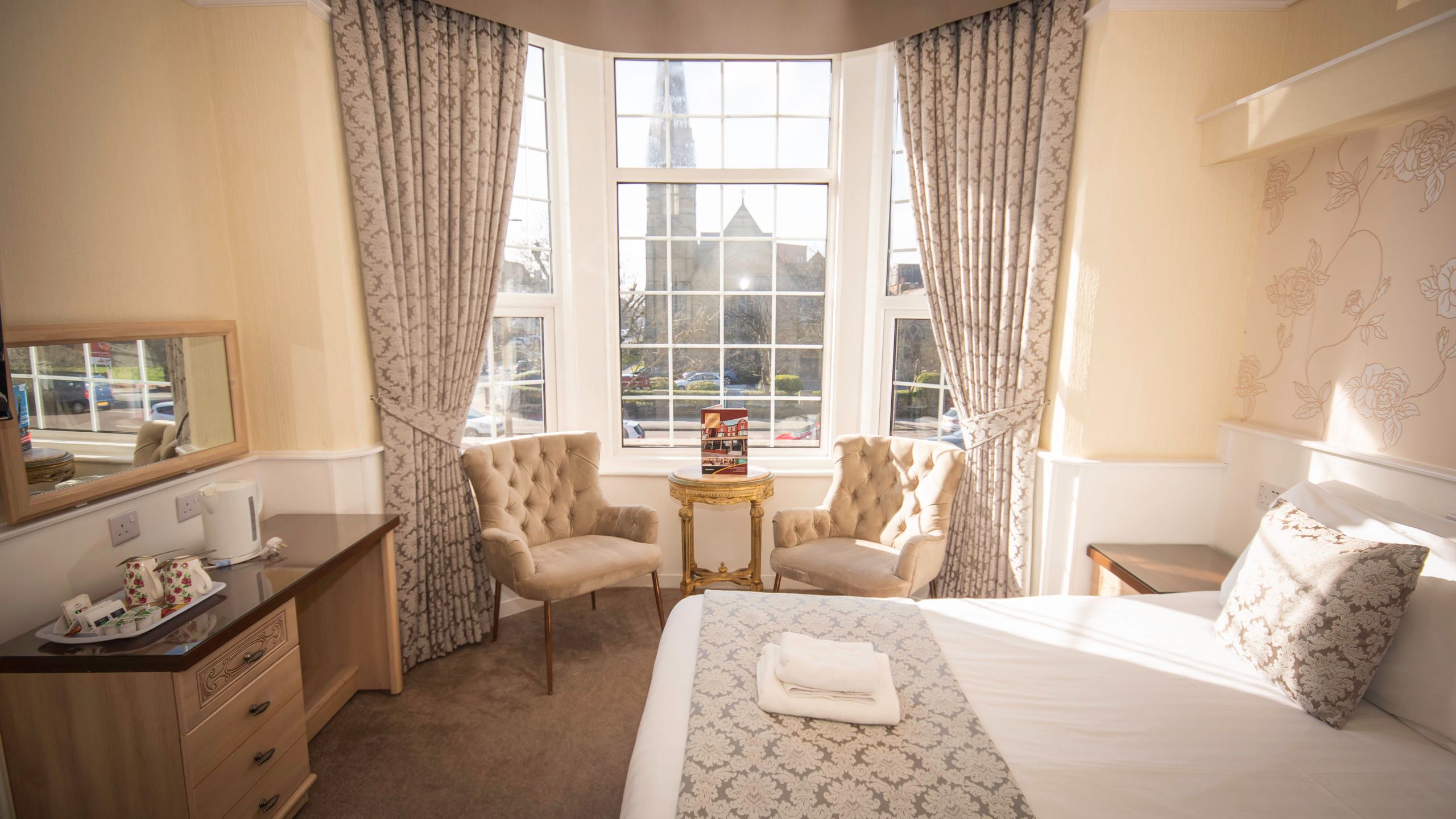 Sunlit hotel bedroom with large bay window, patterned curtains, two armchairs and neatly made bed