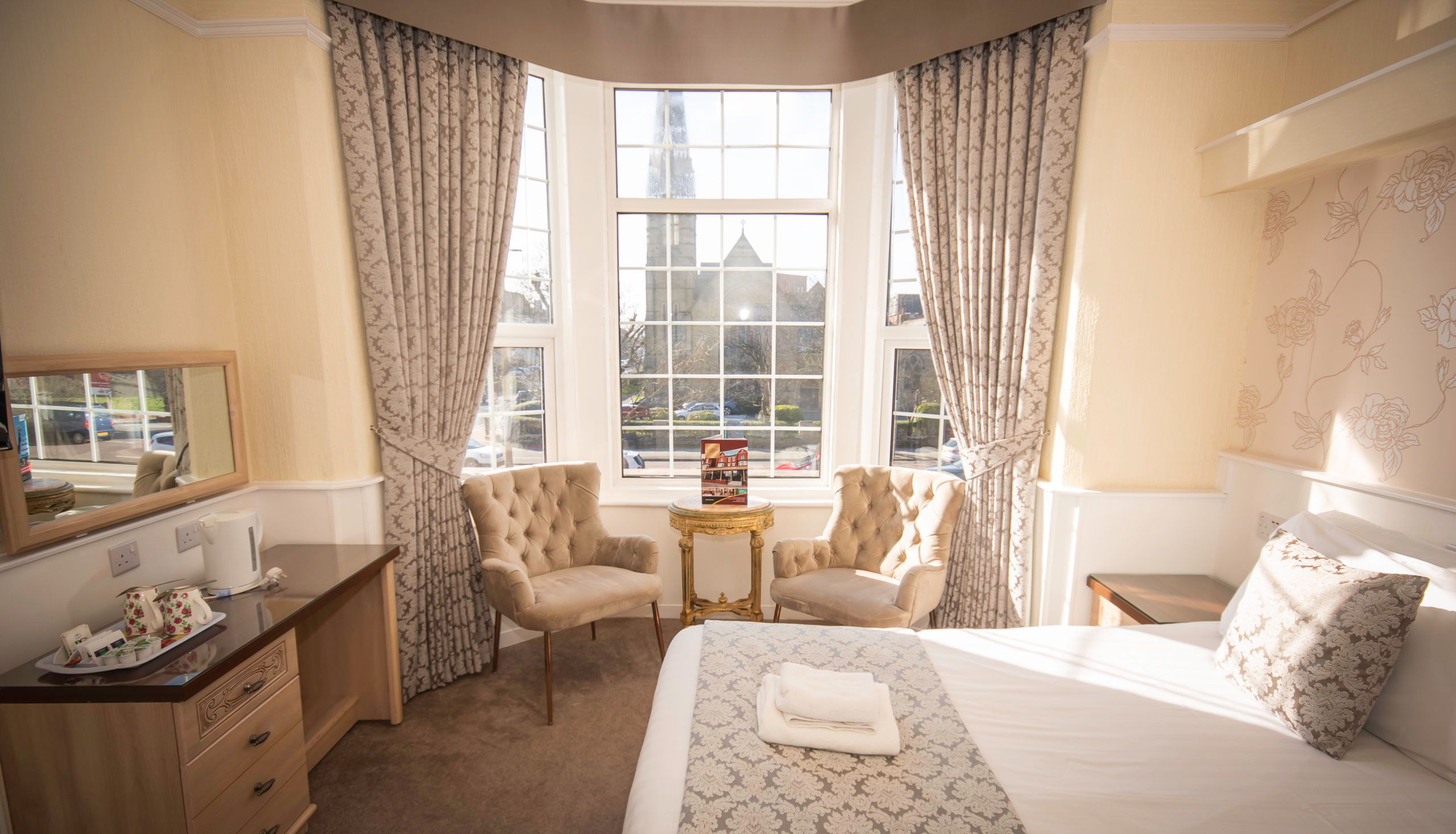 Sunlit hotel bedroom with large bay window, patterned curtains, two armchairs and neatly made bed
