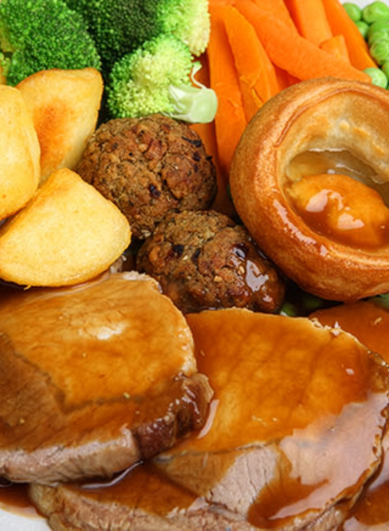 Traditional Sunday roast dinner with sliced meat, roast potatoes, Yorkshire puddings, broccoli, carrots, peas and gravy
