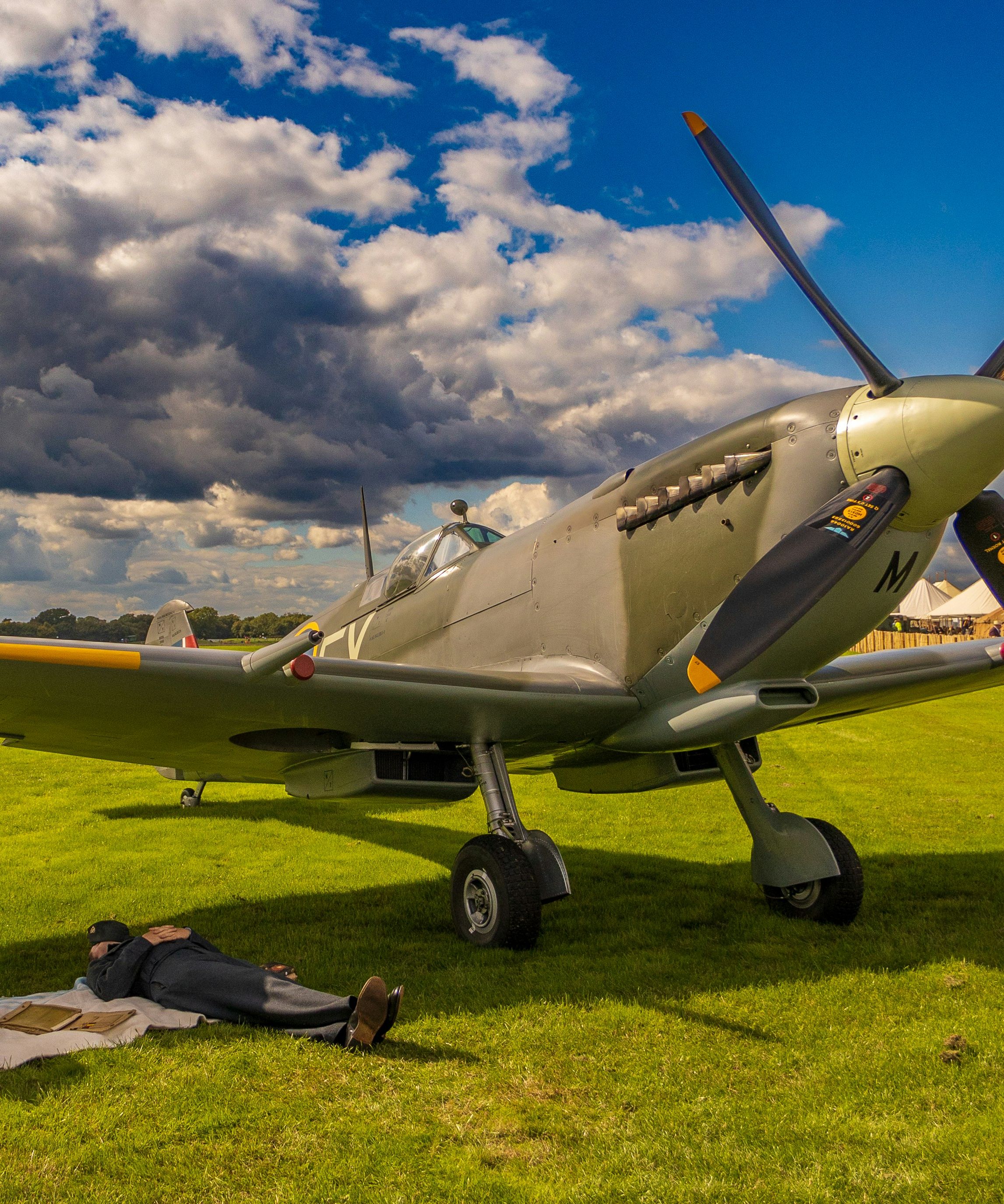 Vintage propeller aircraft on a grassy airfield with people in period clothing and a motorcycle under dramatic clouds