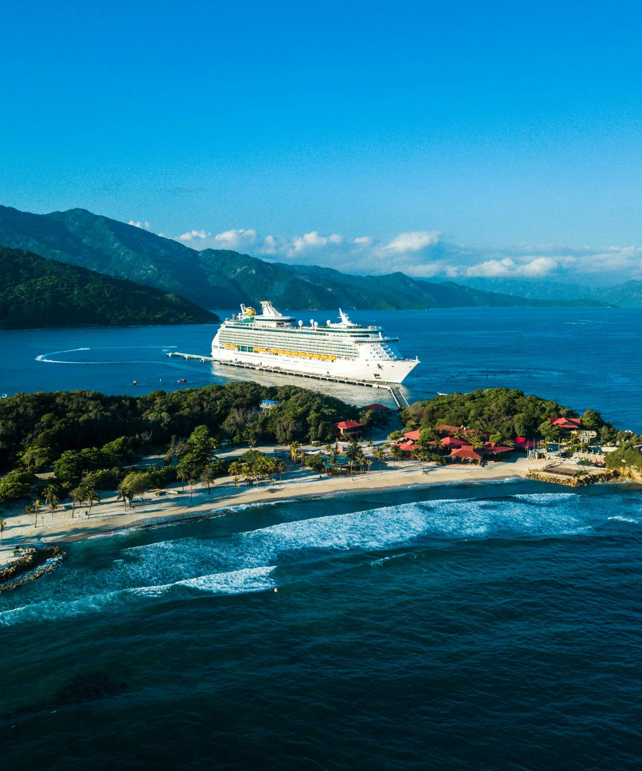 Aerial view of a cruise ship docked by a tropical island with a sandy beach and green mountains