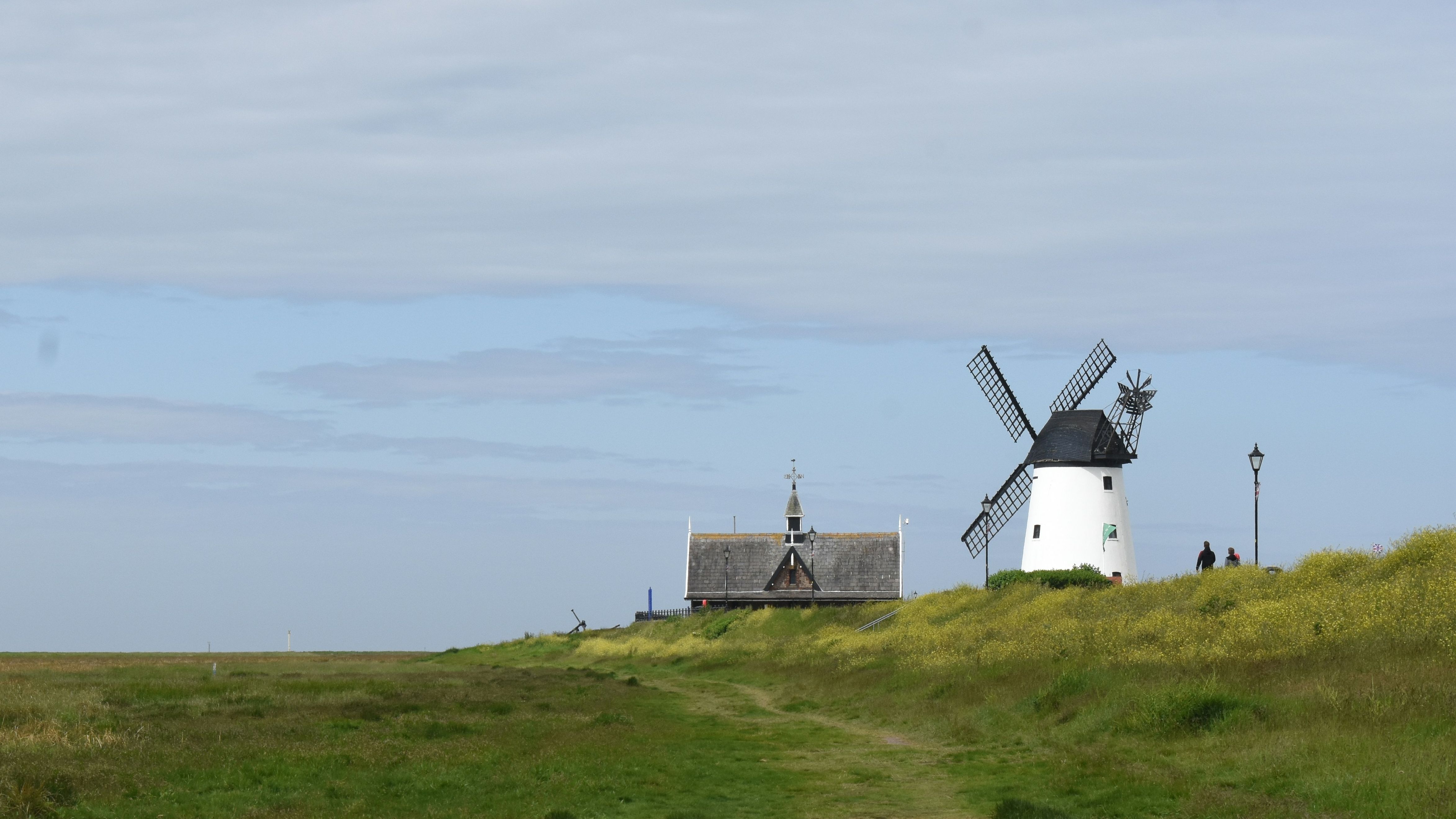 Countryside path leading to a white windmill and cottage on a grassy hill under a cloudy sky