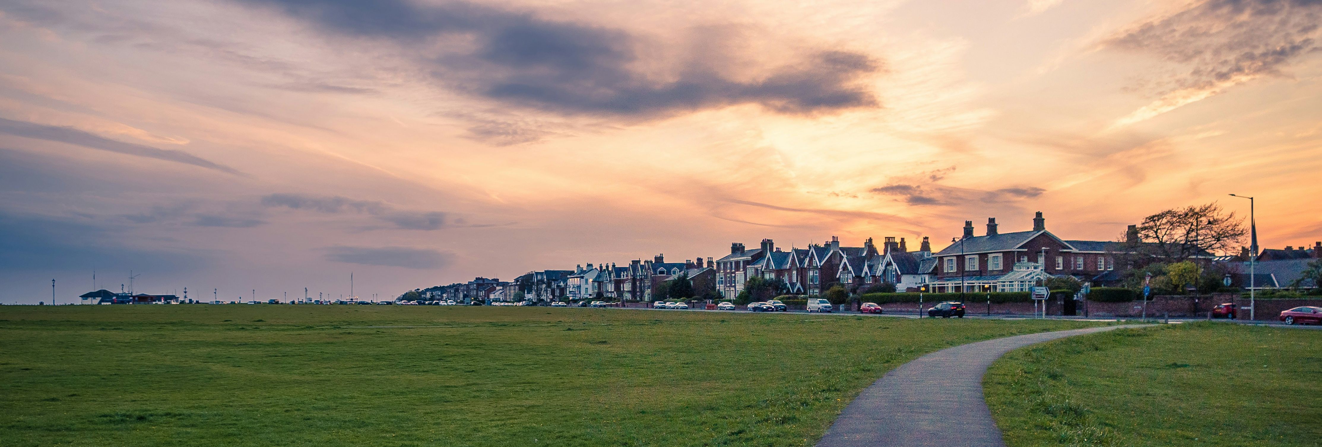 Paved footpath winding through a grassy field towards seaside houses at sunset under dramatic clouds