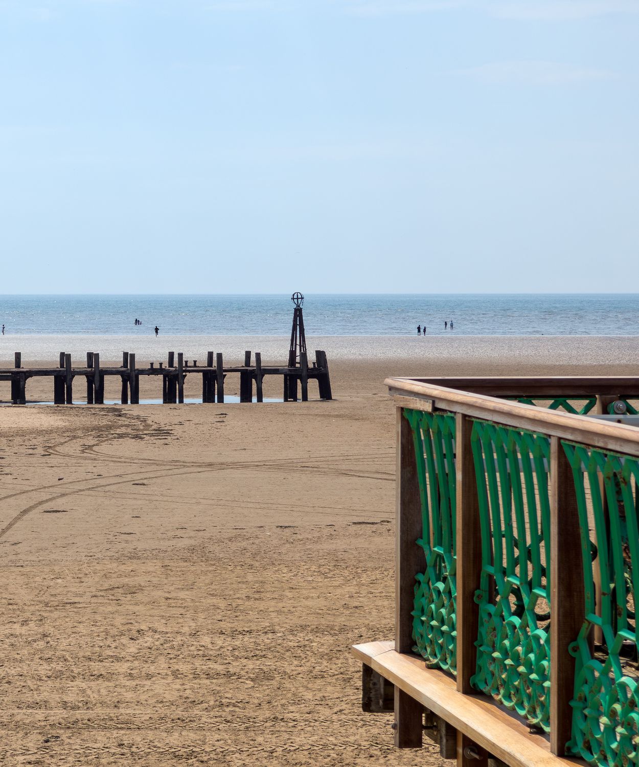 Sandy beach with a wooden groyne at low tide and a green-railed boardwalk in the foreground