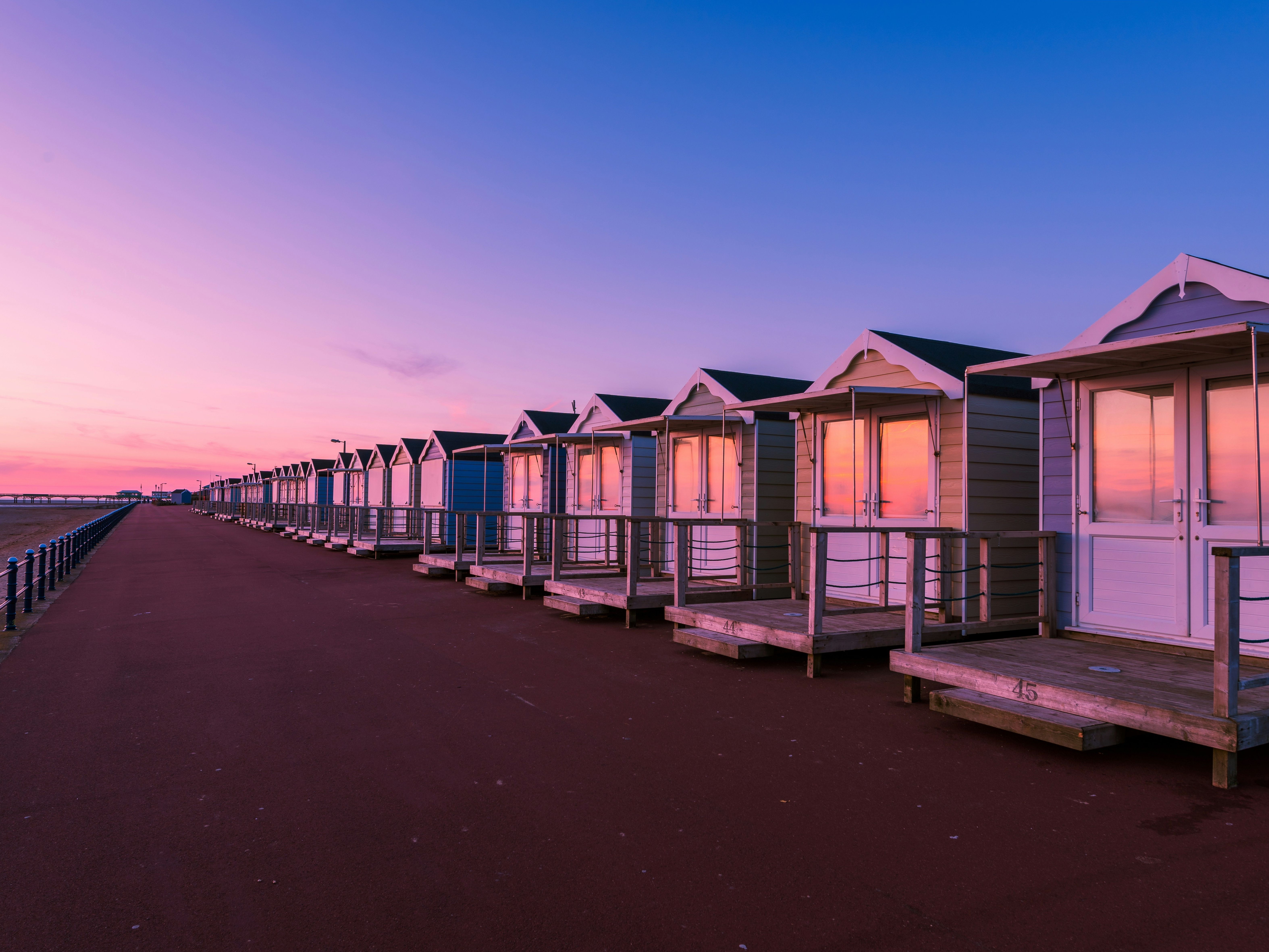 Row of beach huts along a seaside promenade at sunrise, with pink and purple sky reflected in the windows