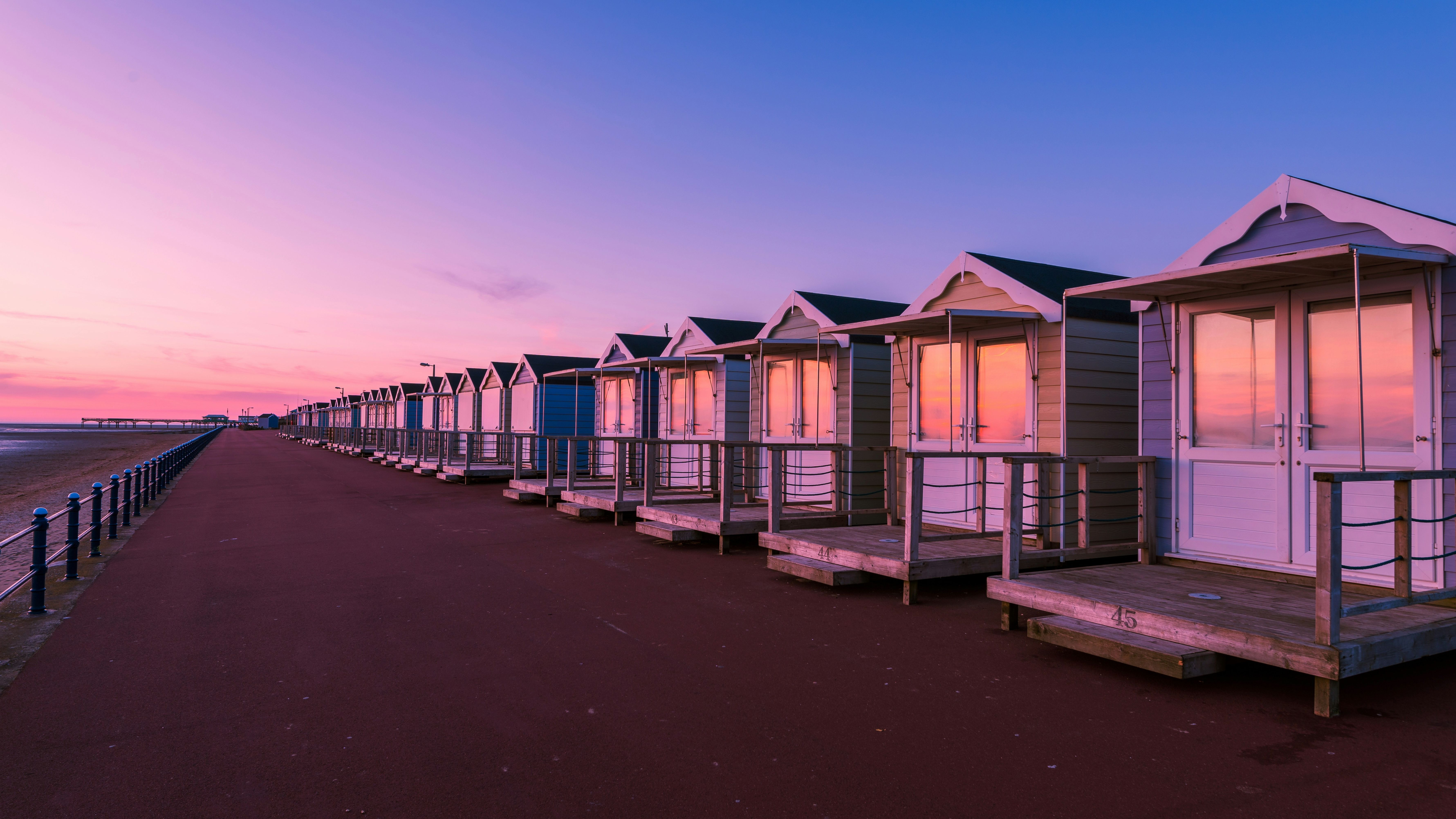 Row of beach huts along a seaside promenade at sunrise, with pink and purple sky reflected in the windows