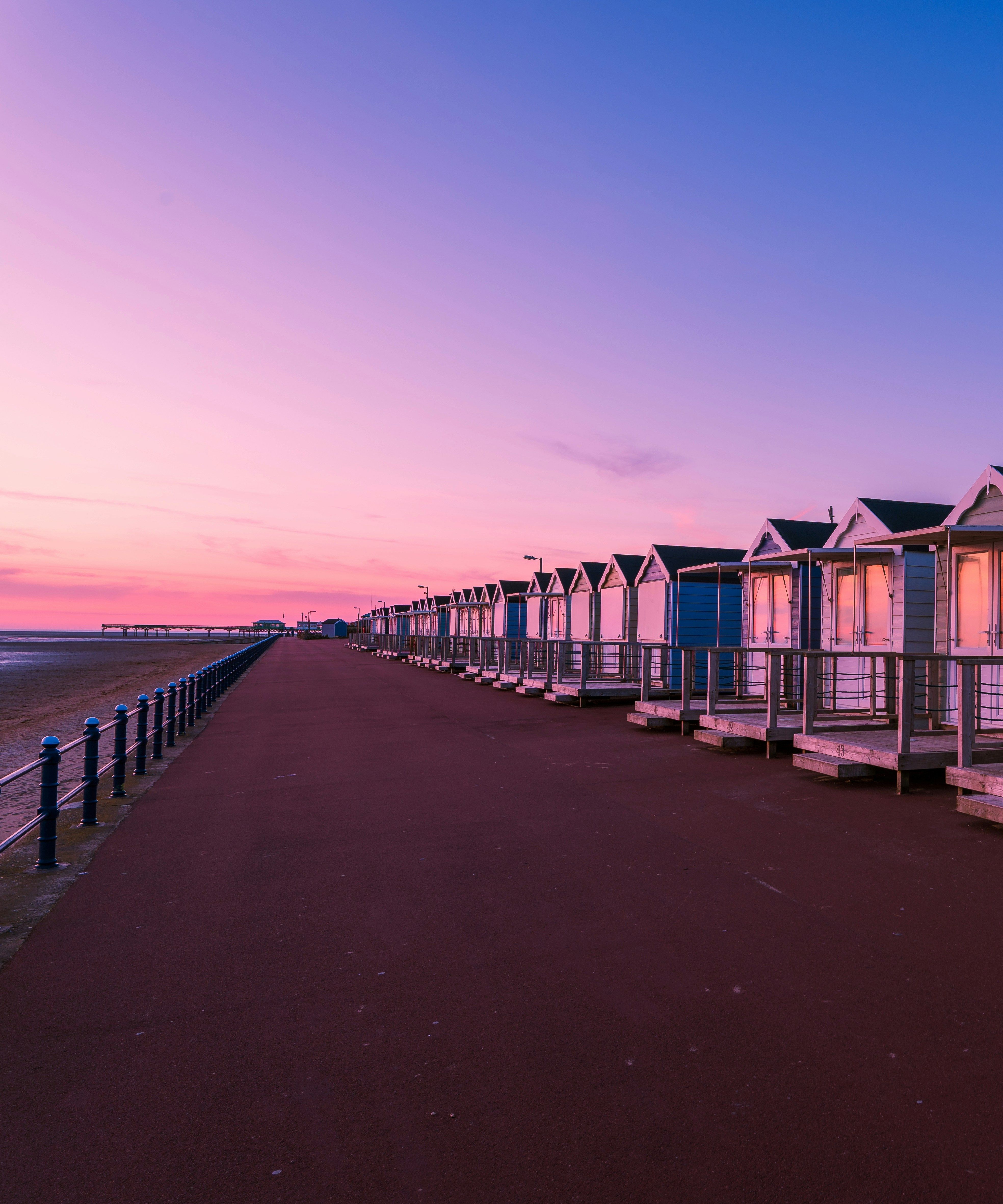 Row of beach huts along a seaside promenade at sunrise, with pink and purple sky reflected in the windows