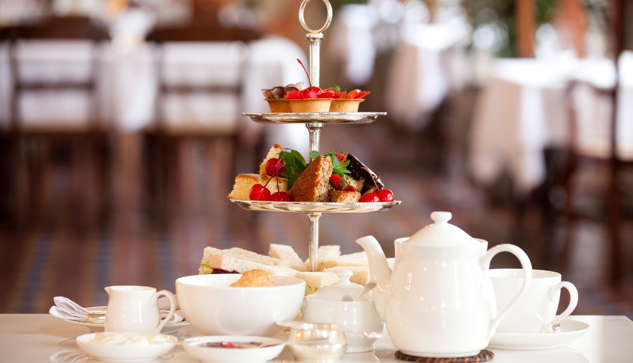 Afternoon tea set with tiered cake stand, sandwiches and fruit pastries beside a white teapot in a restaurant