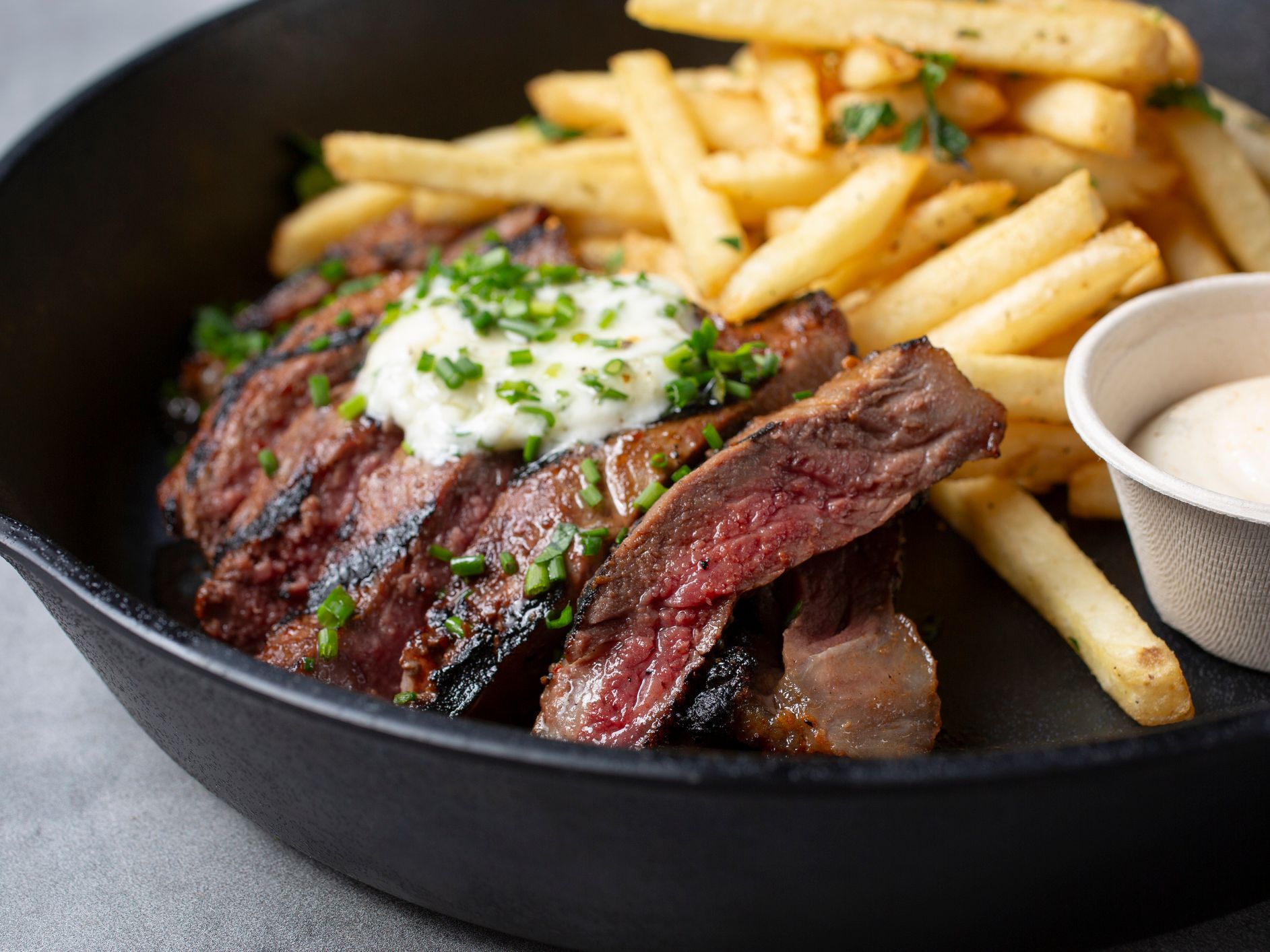 Sliced grilled steak topped with herb butter, served with chips and a small pot of dipping sauce in a black bowl