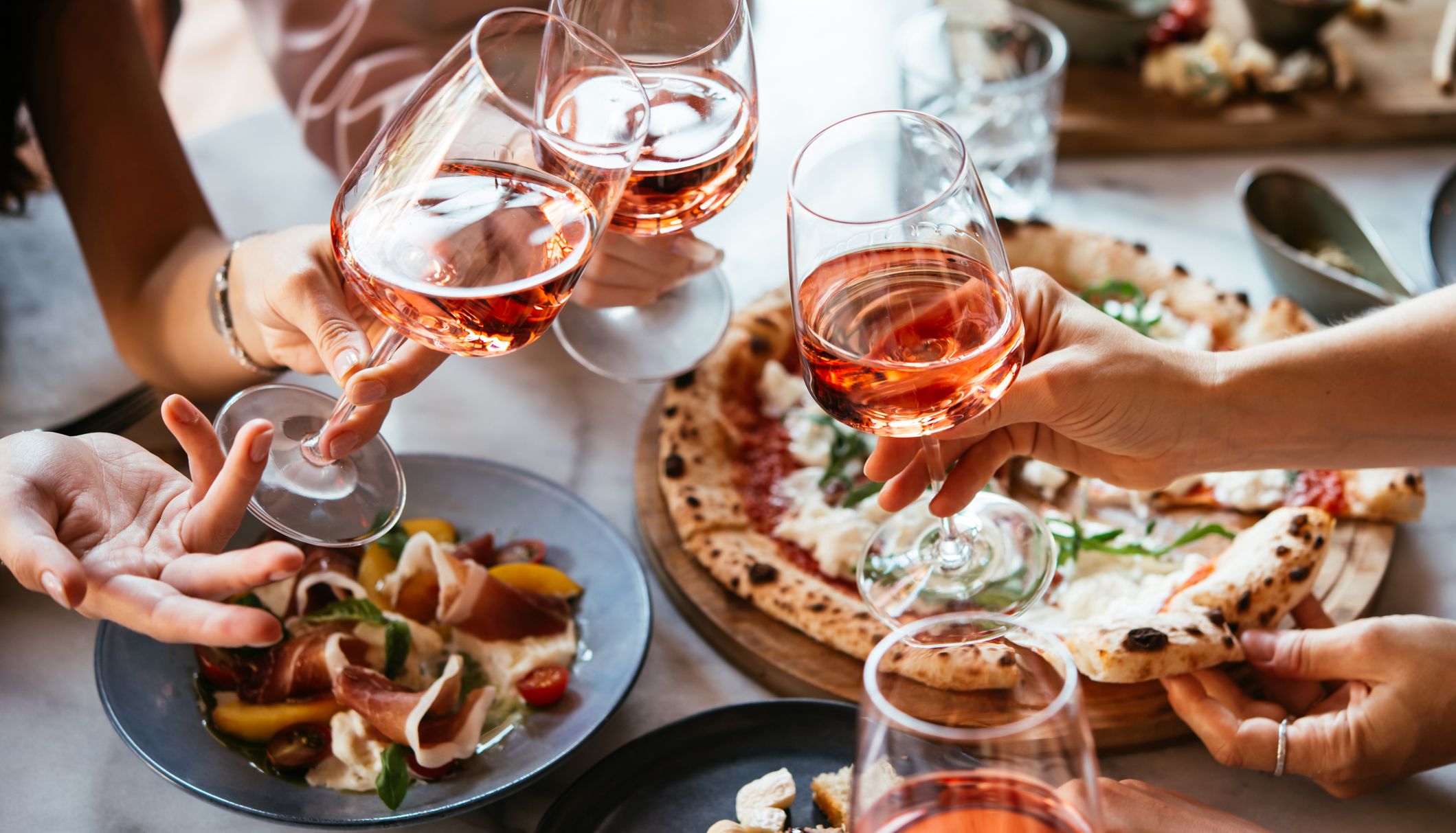 Friends clinking glasses of rosé wine over pizza and antipasti at a restaurant table