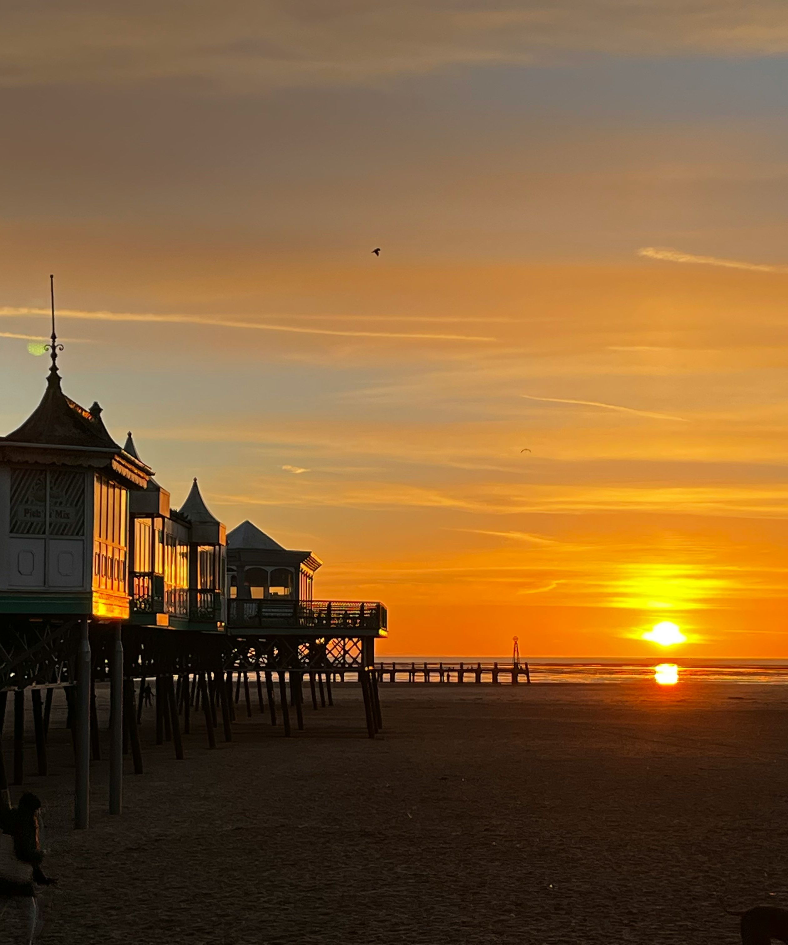 Pier buildings on stilts silhouetted against a golden sunset over a wide sandy beach