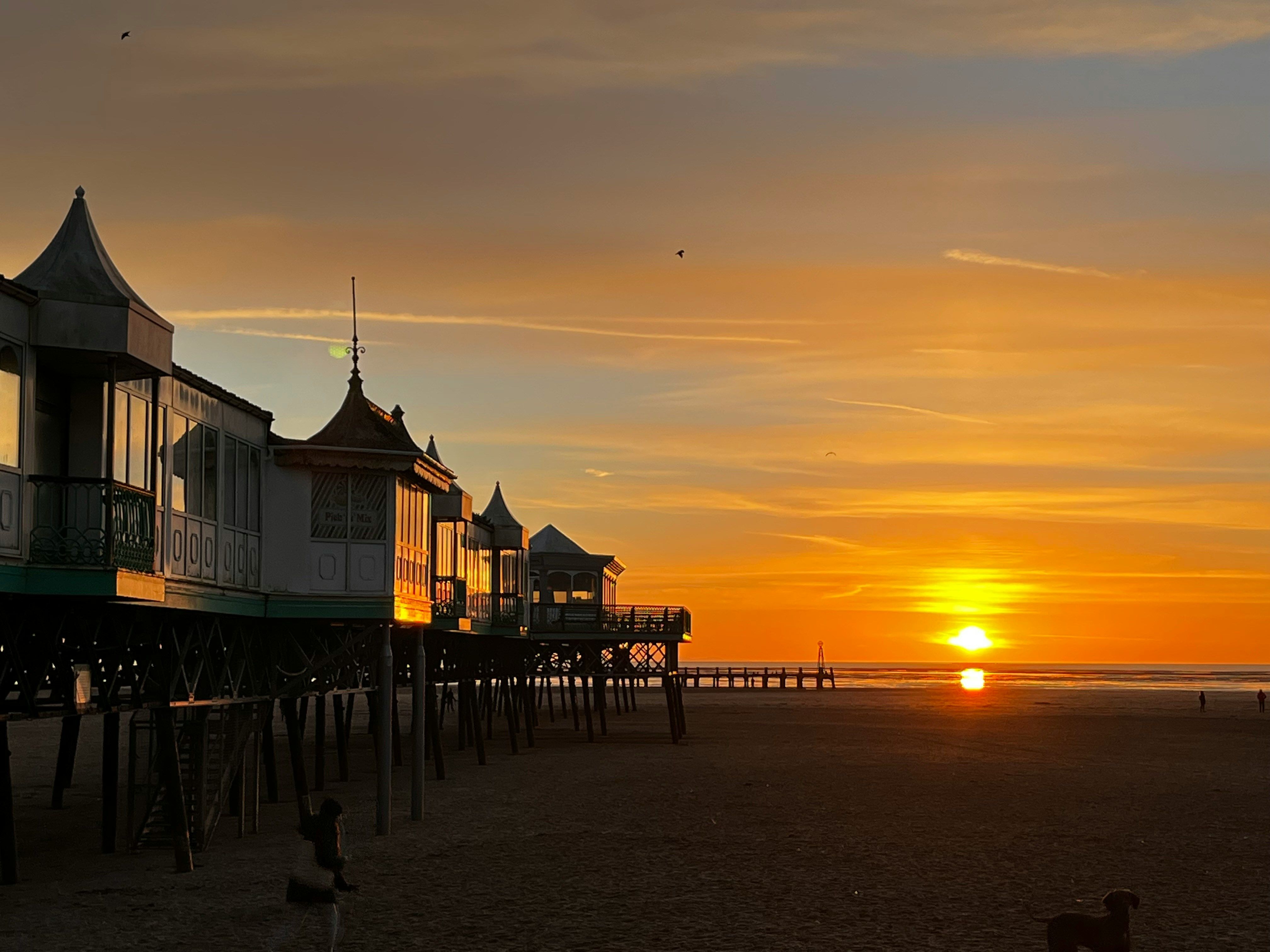 Pier buildings on stilts silhouetted against a golden sunset over a wide sandy beach