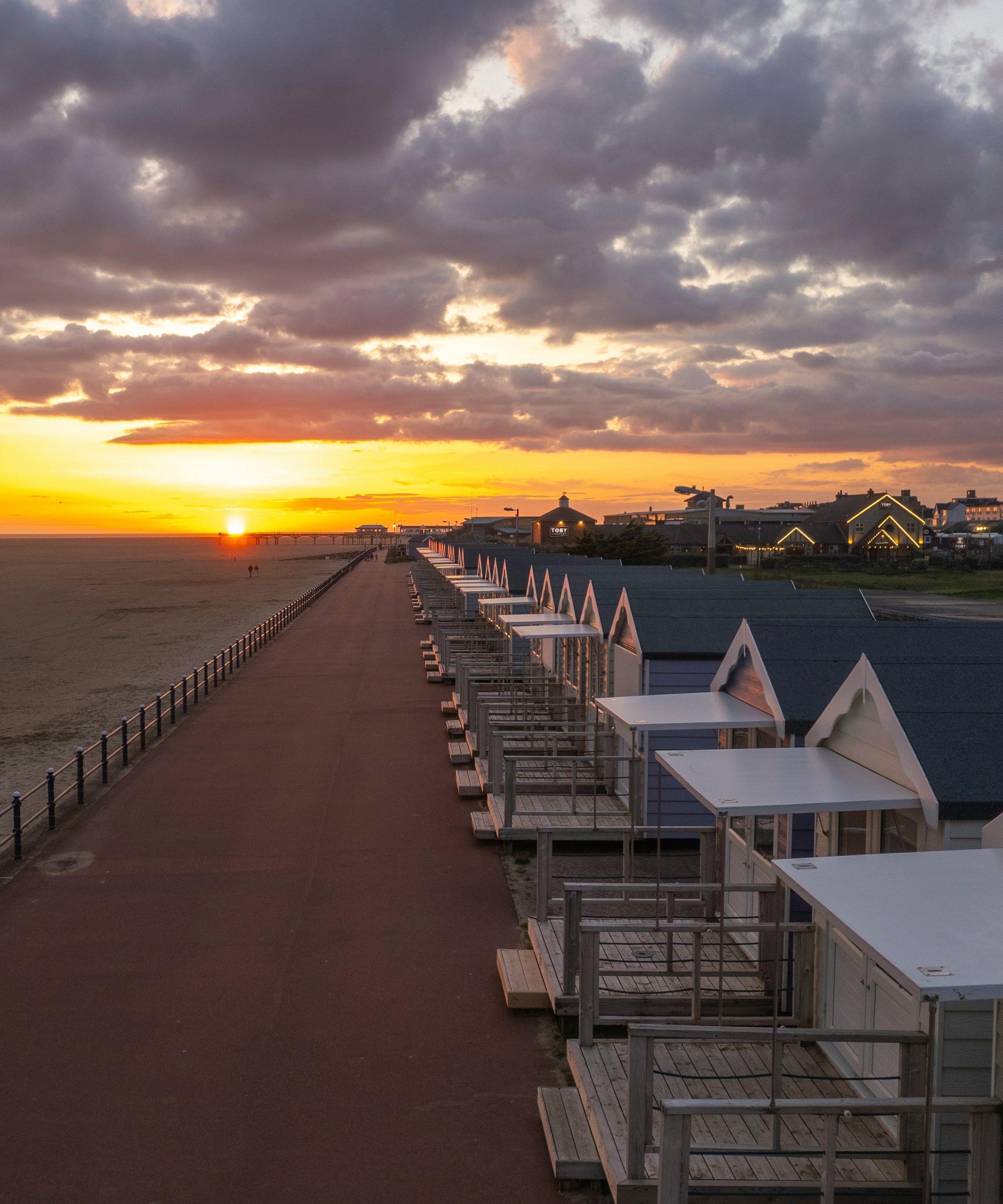 Sunset over a seaside promenade with beach huts beside a wide sandy beach
